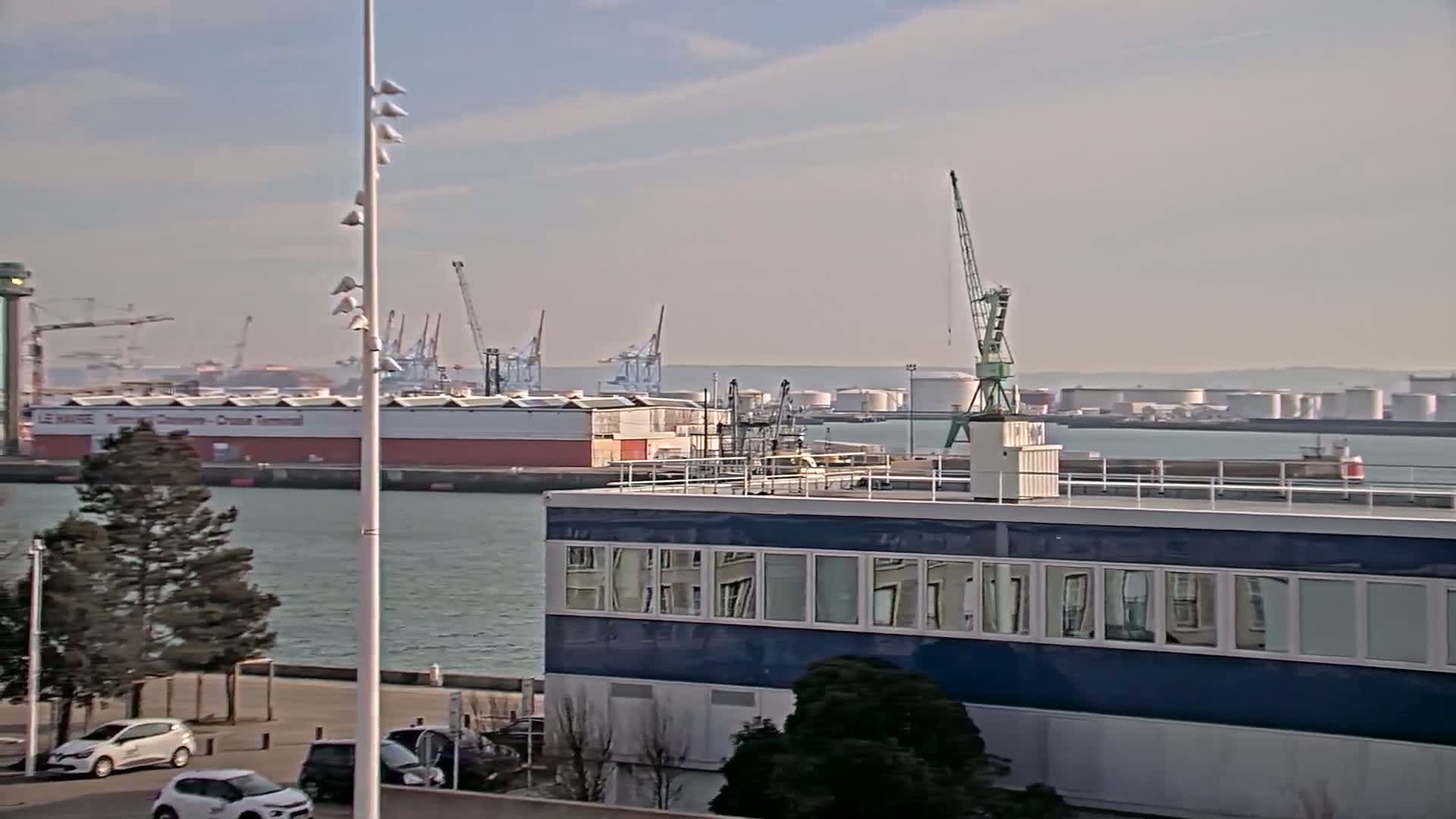 A harbor scene on a hazy day shows several cranes, large cargo ships, and industrial buildings, with cars parked in a lot in the foreground.