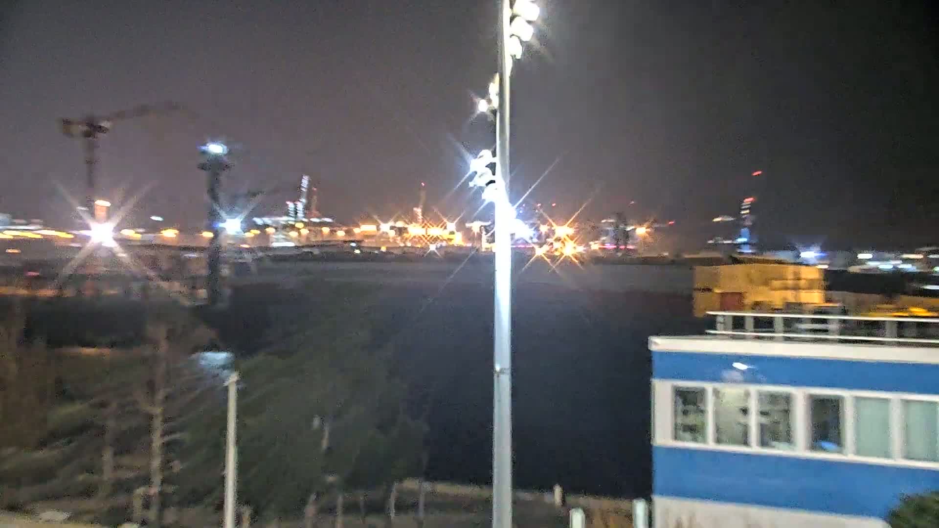 A nighttime view of a brightly lit industrial area across a body of water, featuring a tall light post in the foreground and a small blue building to the right.