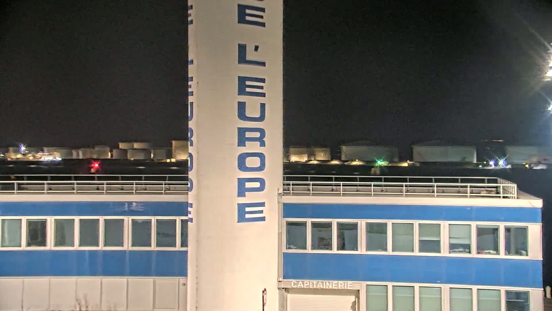 A tall white pillar with blue lettering stands in front of a blue building at night, with a row of industrial tanks visible in the background.