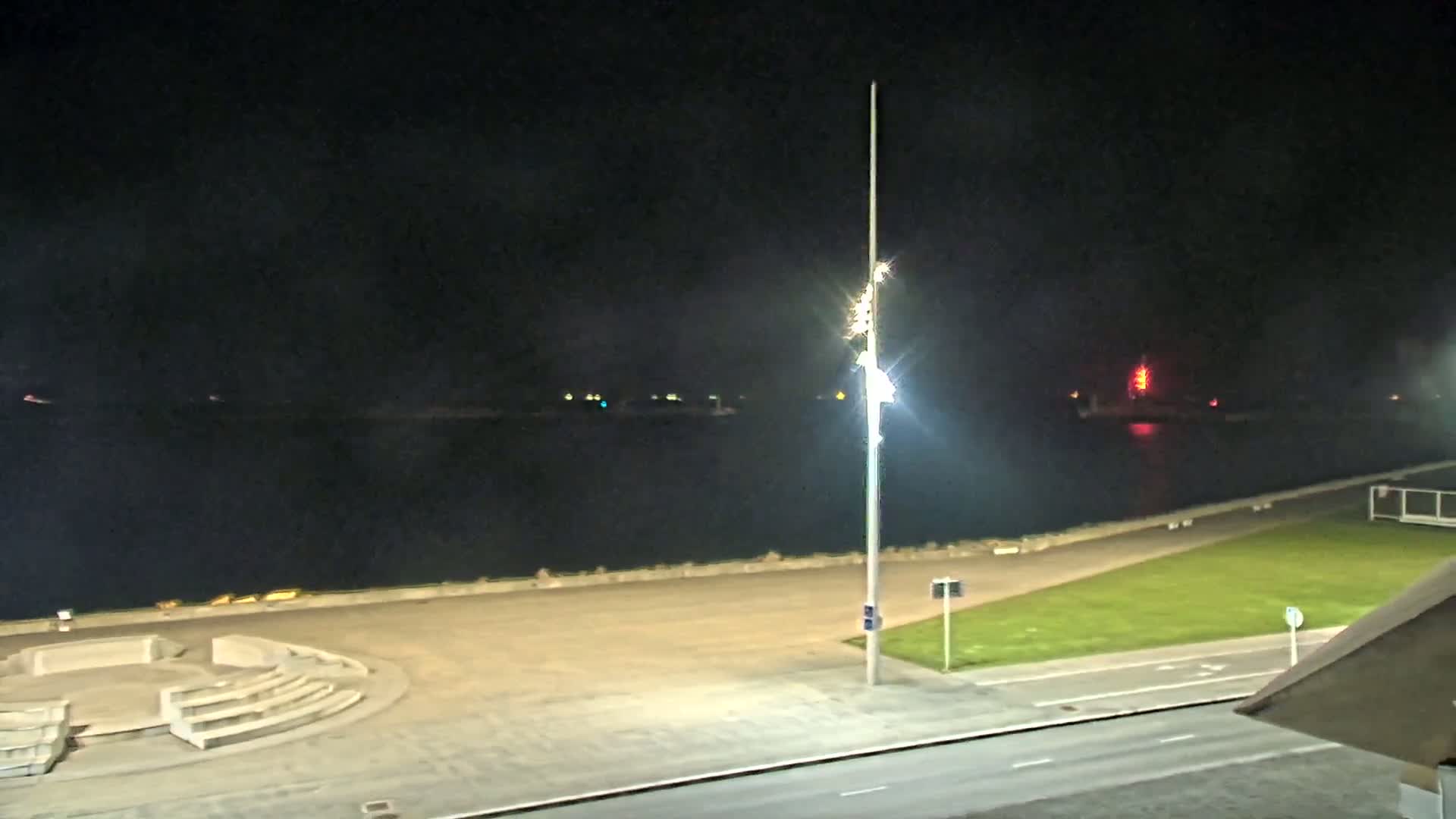 A nighttime view of a waterfront area with a paved plaza, a grassy area, a road, and a tall light pole, overlooking calm dark water with distant lights.