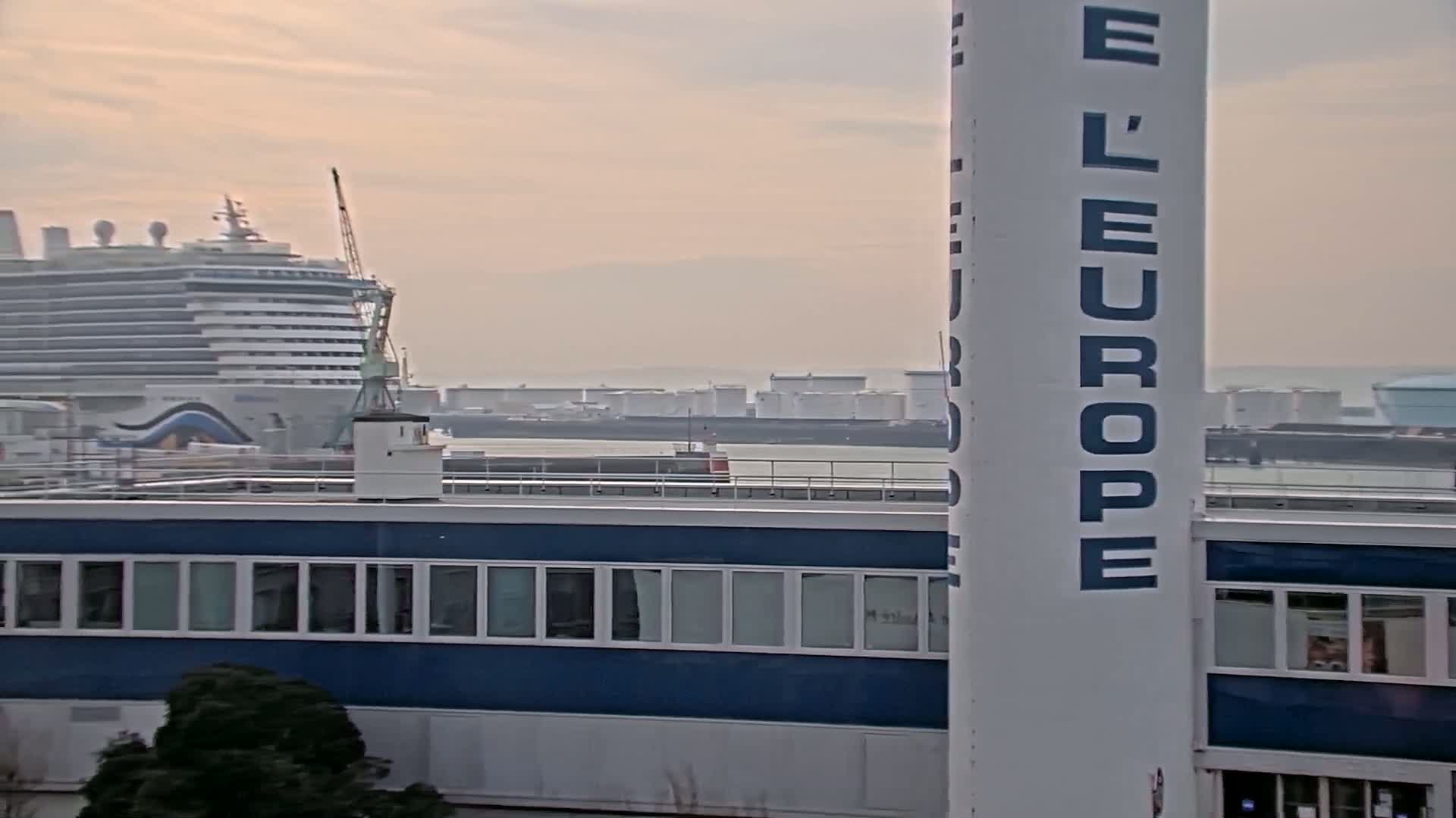 A large cruise ship is docked near a building with many windows, under a hazy sky; in the distance, there are industrial storage tanks.