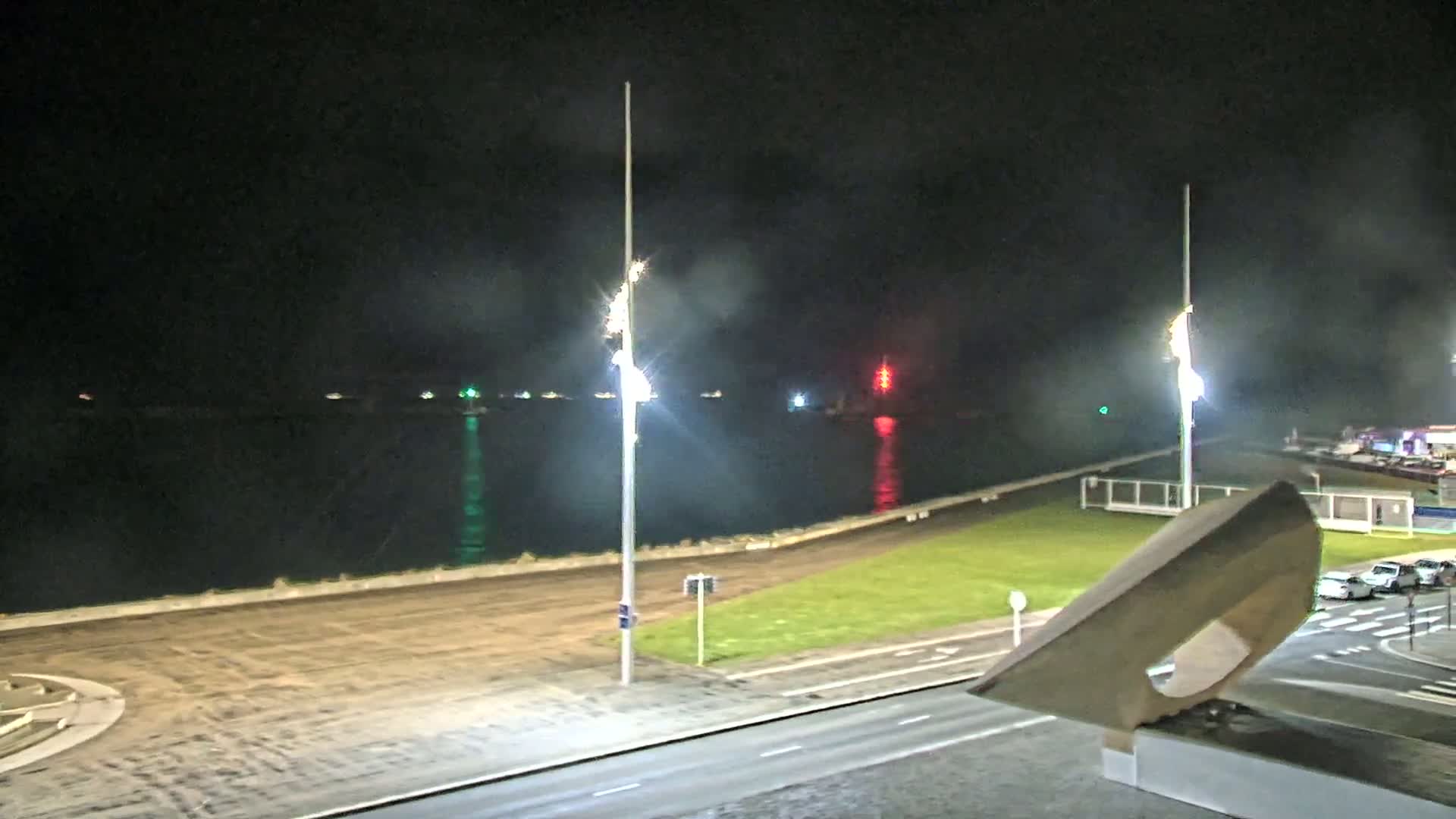 A nighttime view of a calm body of water with lights on the far shore, a road and grassy area in the foreground, and tall light posts illuminating the scene.