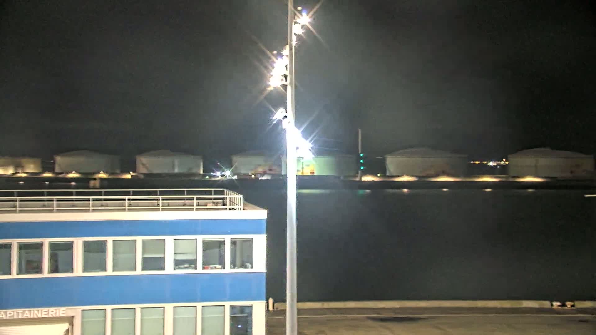 A brightly lit tall pole stands beside a blue building at night near a body of dark water with several large, illuminated storage tanks in the background.