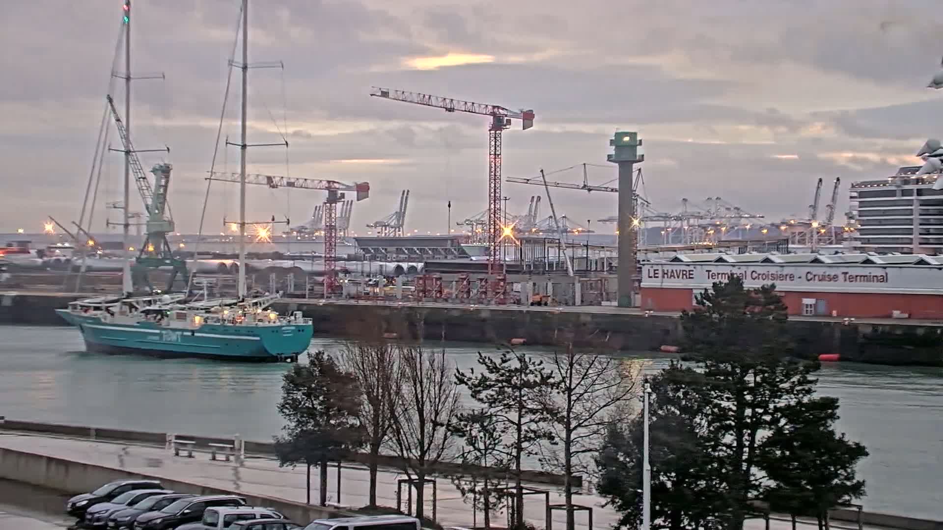 A teal-hulled sailing ship is in a harbor under a cloudy sky, with construction cranes and other ships in the background.