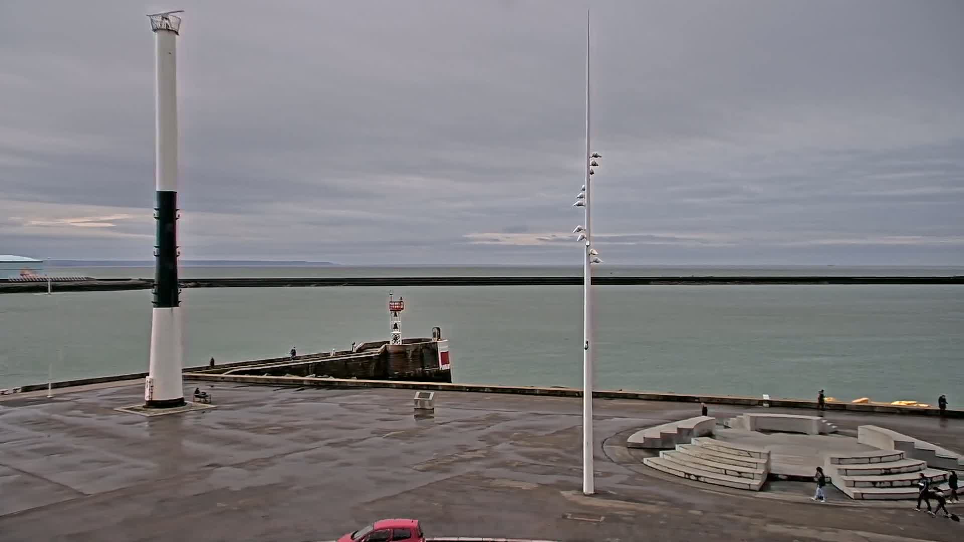 A tall lighthouse stands on a wet, empty plaza overlooking a calm sea under a cloudy sky.