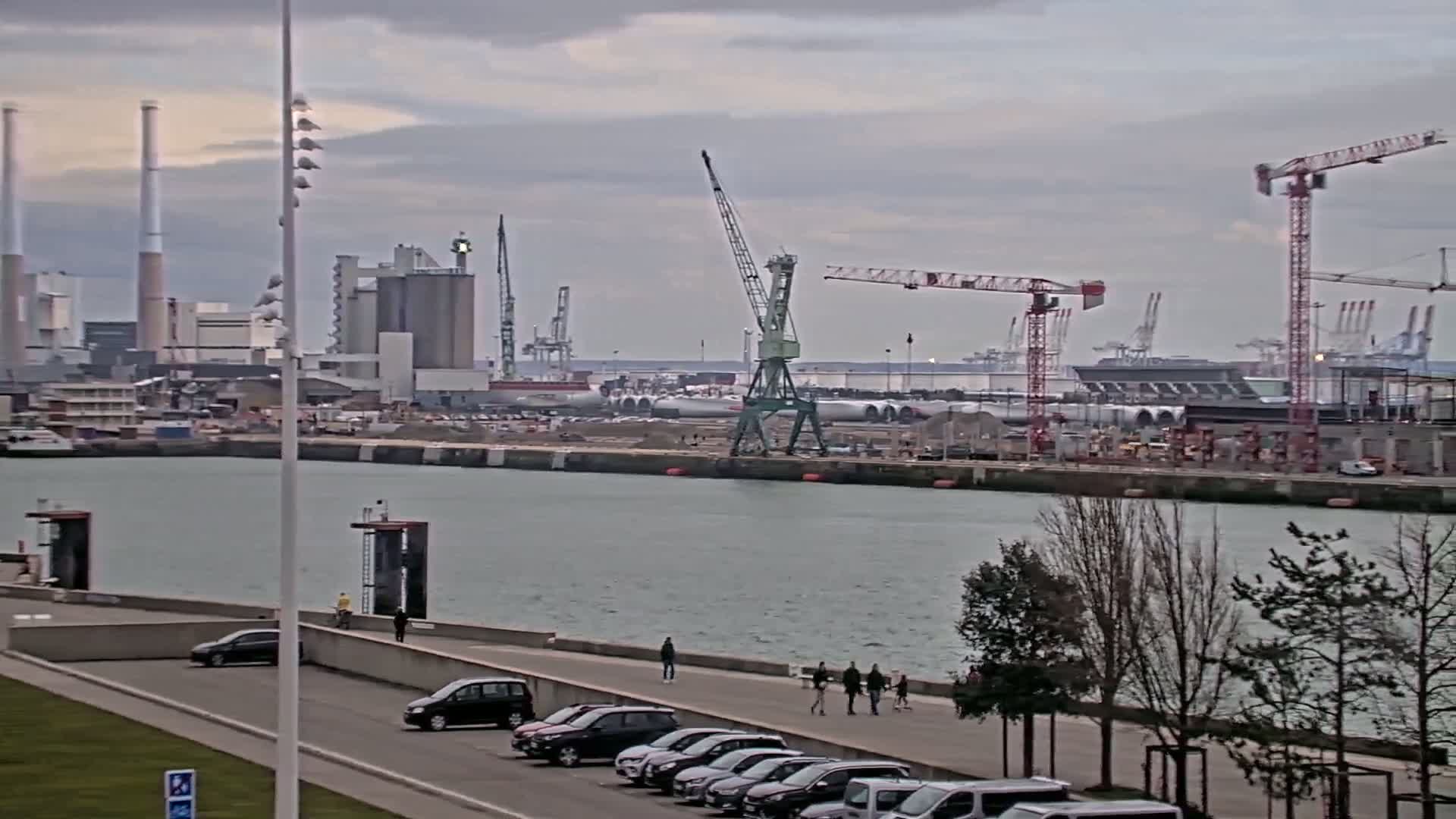 A waterfront view shows a harbor with industrial buildings, cranes, and several parked cars along a walkway on an overcast day.