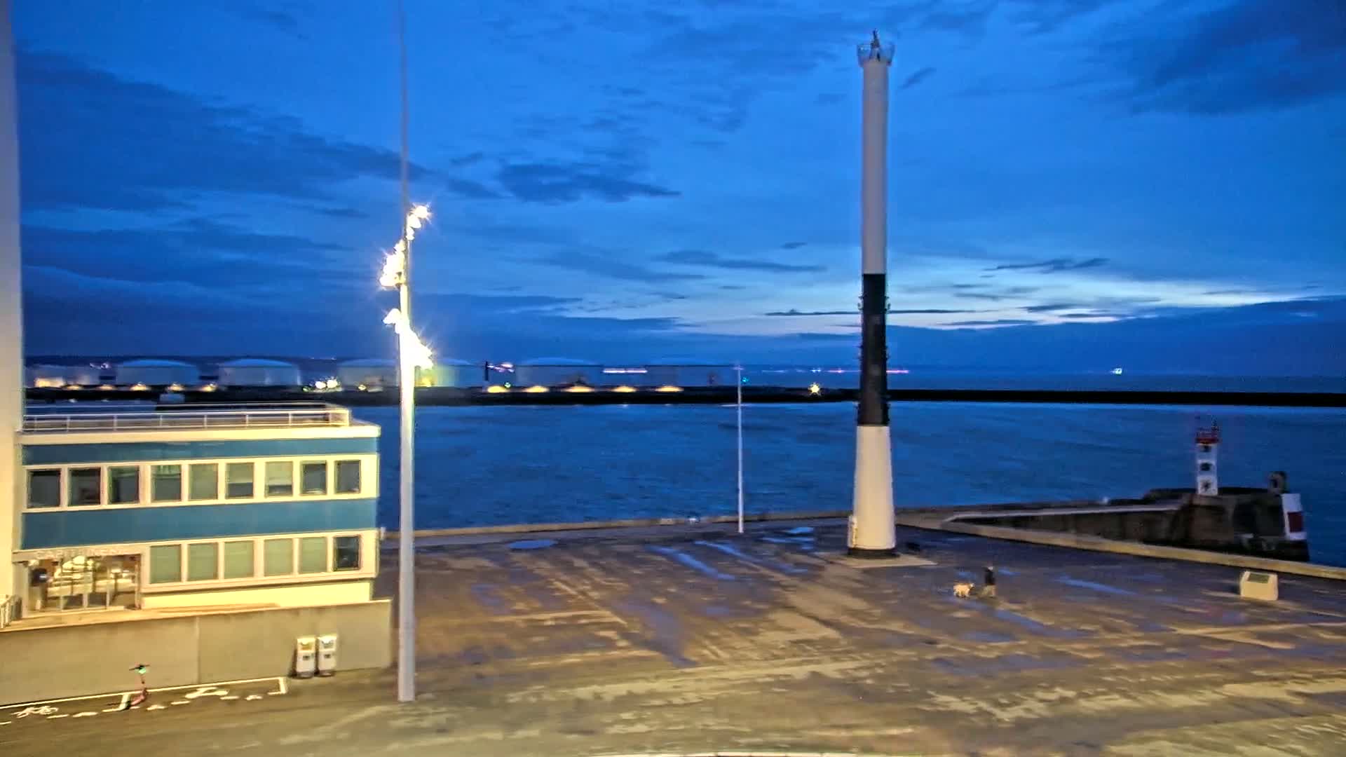 A person walks a dog on a dark, empty dock at twilight next to a tall, black and white lighthouse, overlooking a calm body of water with industrial structures in the background.