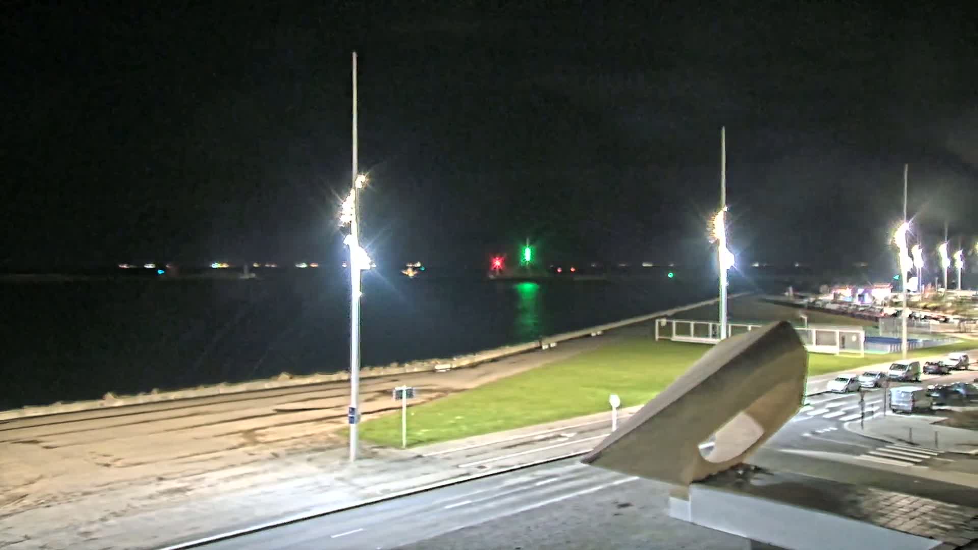 A nighttime view of a waterfront area with a calm body of water, streetlights illuminating a roadway and grassy area next to a seawall, and parked cars visible in the distance.