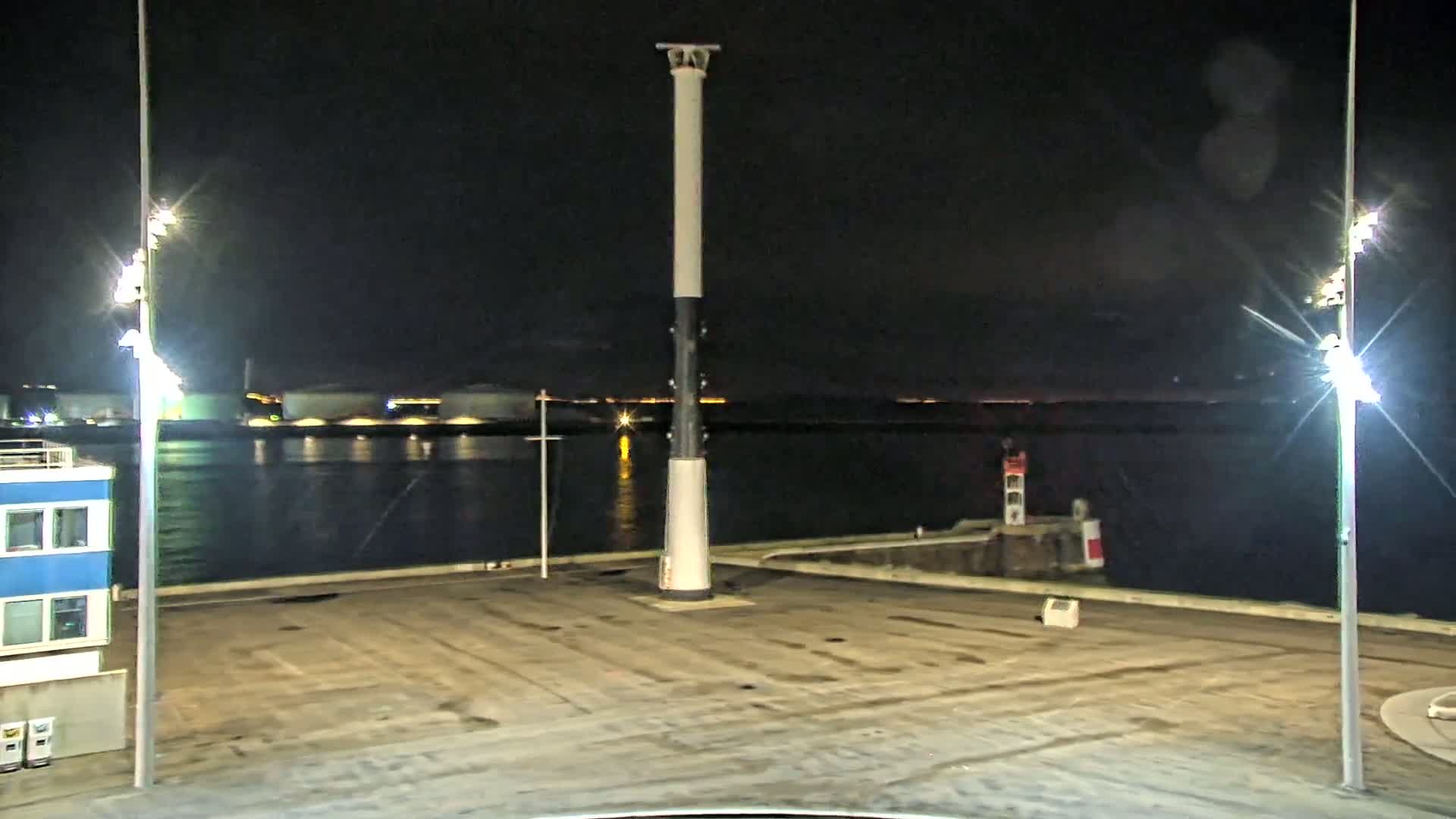 A tall, slender lighthouse-like structure stands in a dark, empty port area at night, illuminated by bright lights against a calm, dark body of water.
