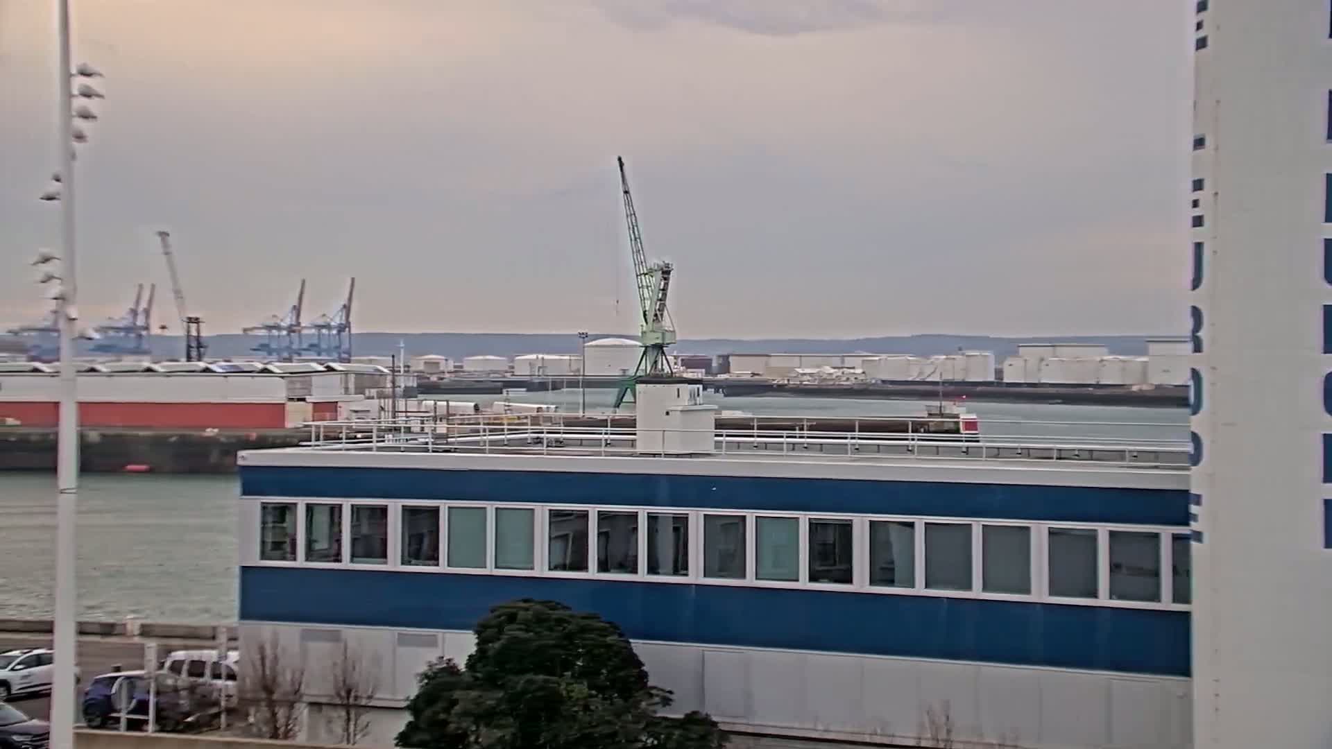 A blue building with many windows sits near a body of water with a crane and distant industrial structures under an overcast sky.