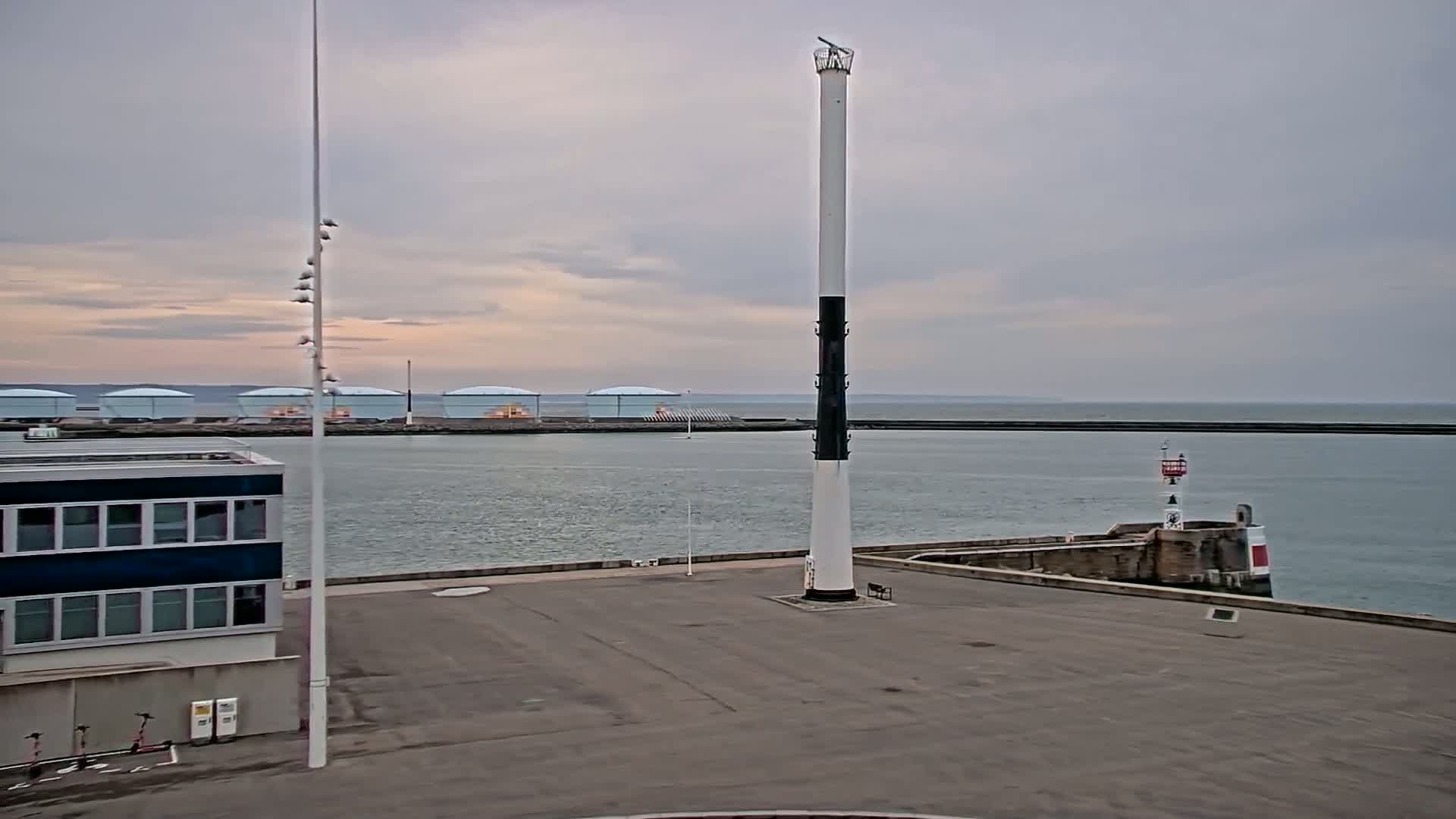 A tall, white and black lighthouse stands in a harbor on an overcast day, with several large storage tanks visible in the distance.