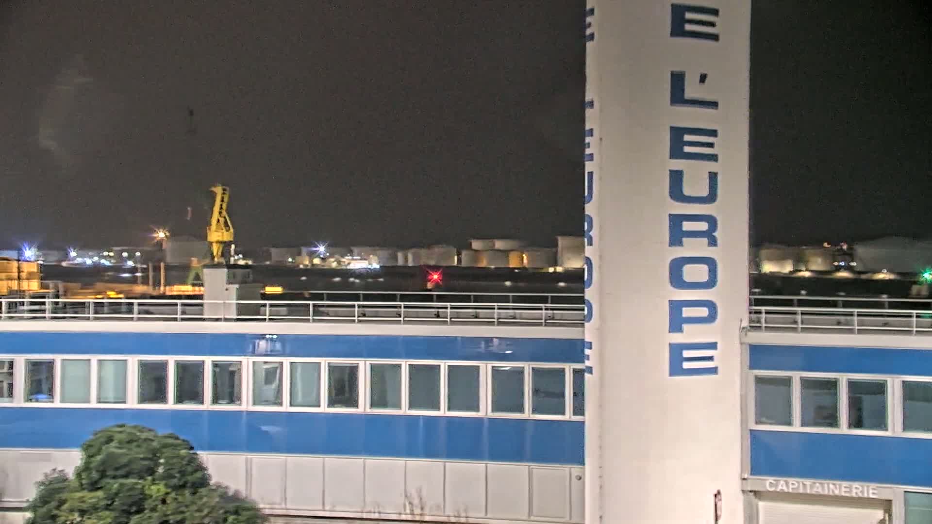 A nighttime view of a blue building with a tall, white pillar displaying "L'EUROPE" and a crane visible in the distance under a dark sky.