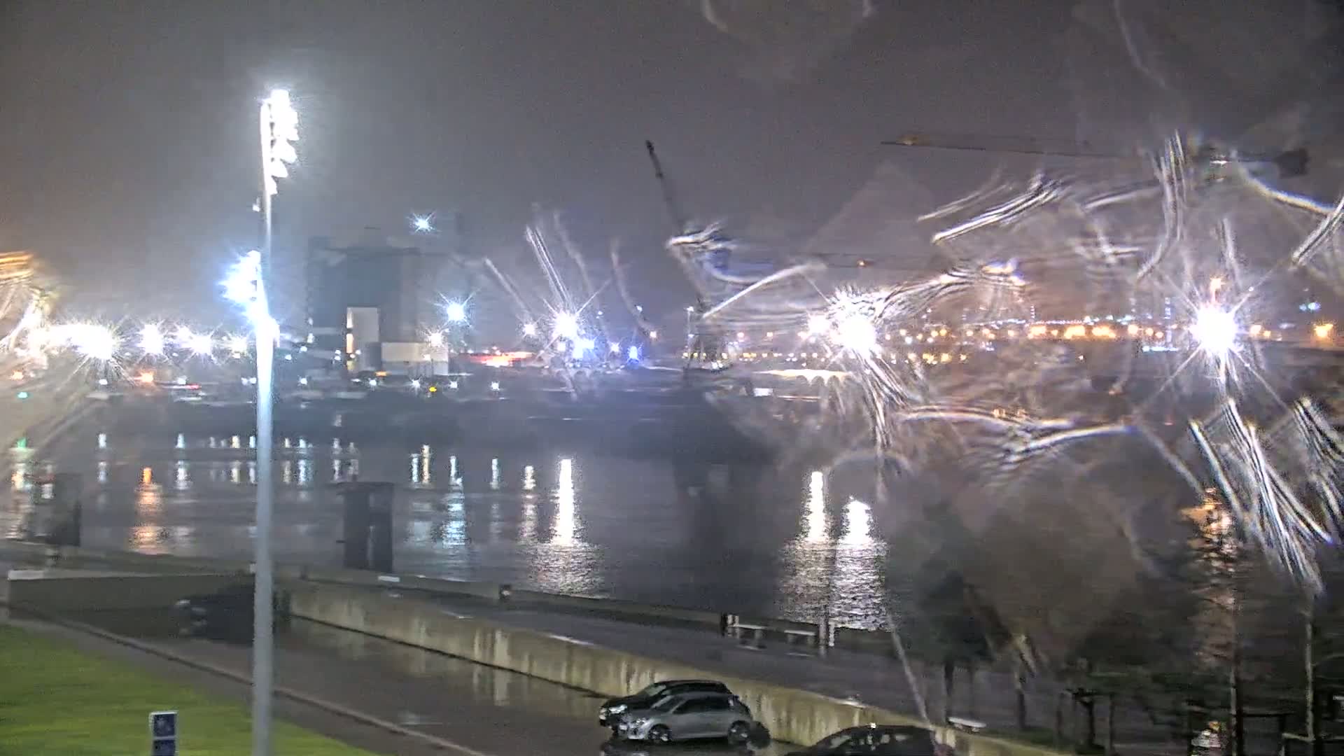 A nighttime view of a rain-slicked waterfront with city lights reflecting on the water, cars parked nearby, and a tall light post in the foreground.