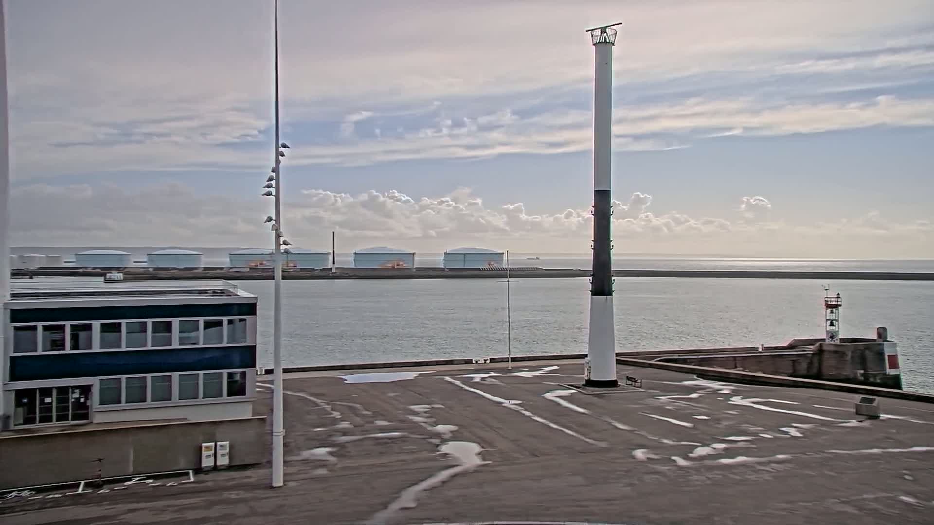 A harbor scene under a partly cloudy sky shows a tall, slender lighthouse next to a building with many windows, with several large storage tanks visible in the distance across the water.