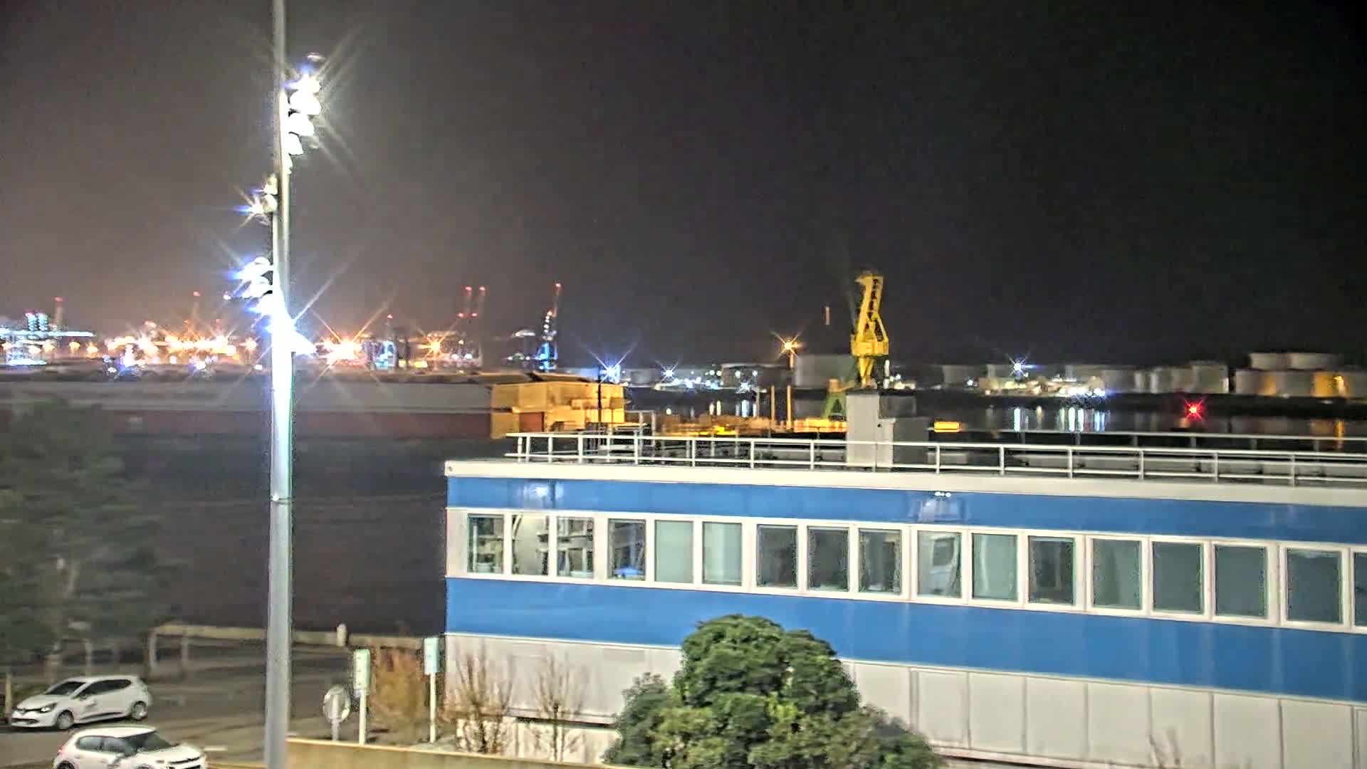 A brightly lit industrial port at night, featuring a blue building in the foreground and a crane in the background.