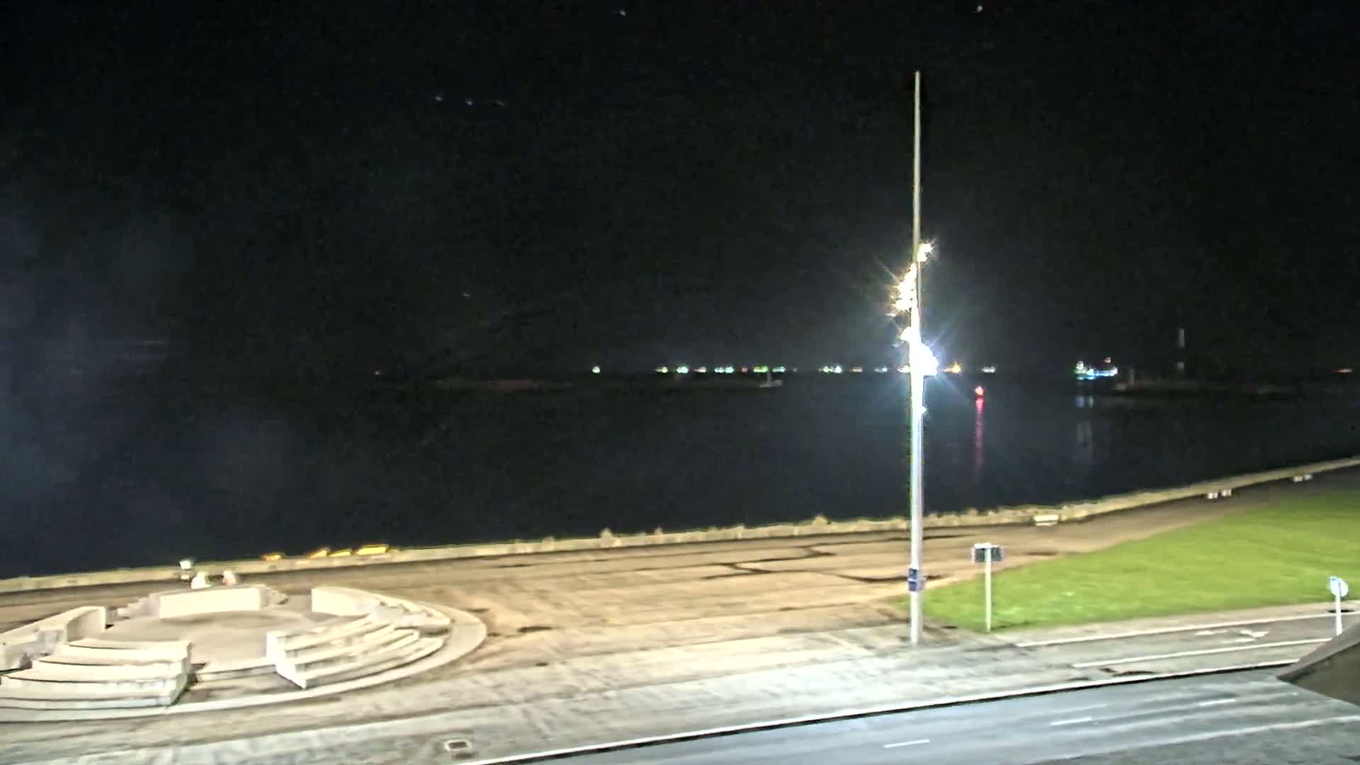 A nighttime view of a calm body of water with a distant shoreline showing lights, and a paved area with a circular structure in the foreground.
