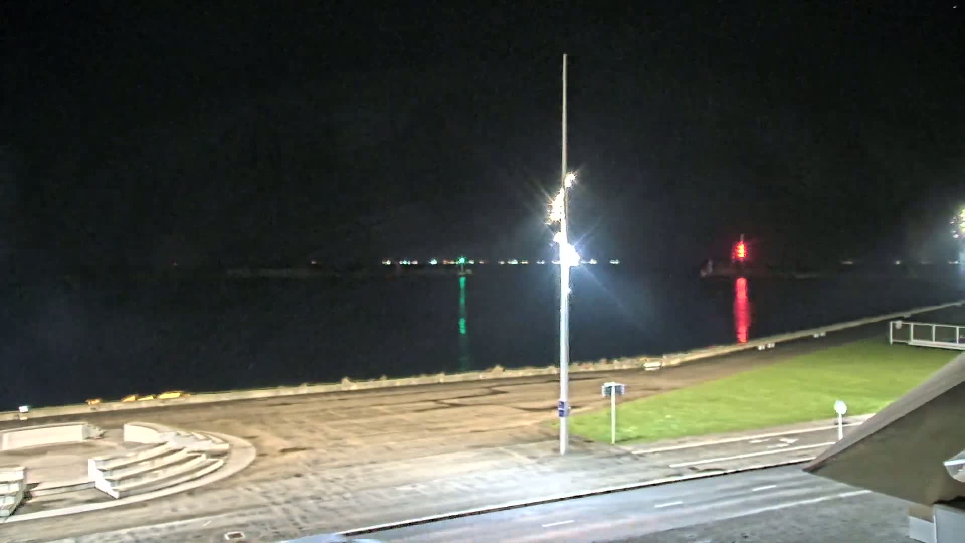 A nighttime view of a waterfront area with a calm body of water, a paved area with a seating structure, and a grassy patch, all illuminated by artificial lighting.