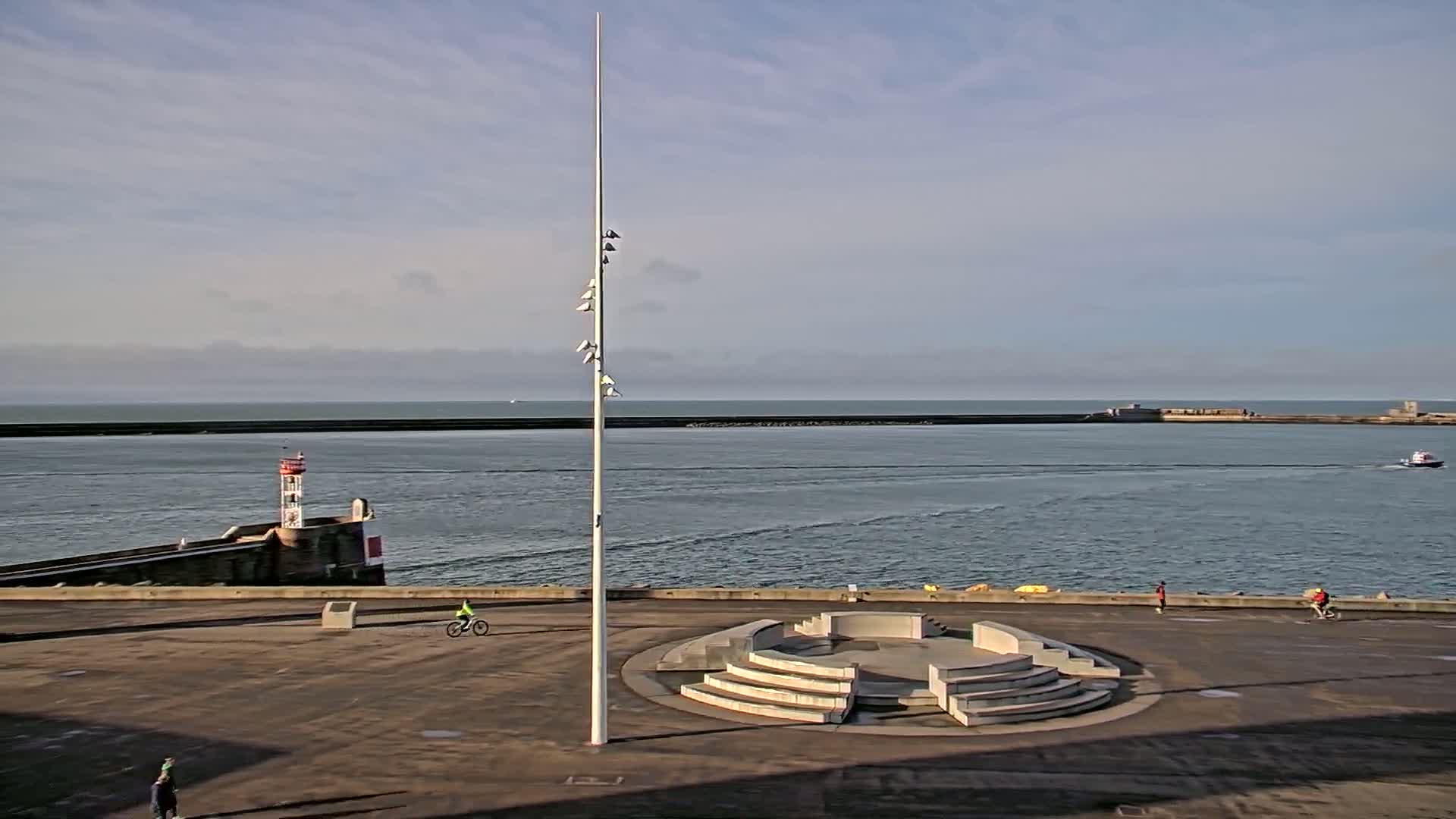 A mostly sunny day shows a harbor with a lighthouse and a tall flagpole at the center of an empty paved plaza featuring a circular tiered seating area, with several people walking and biking nearby.