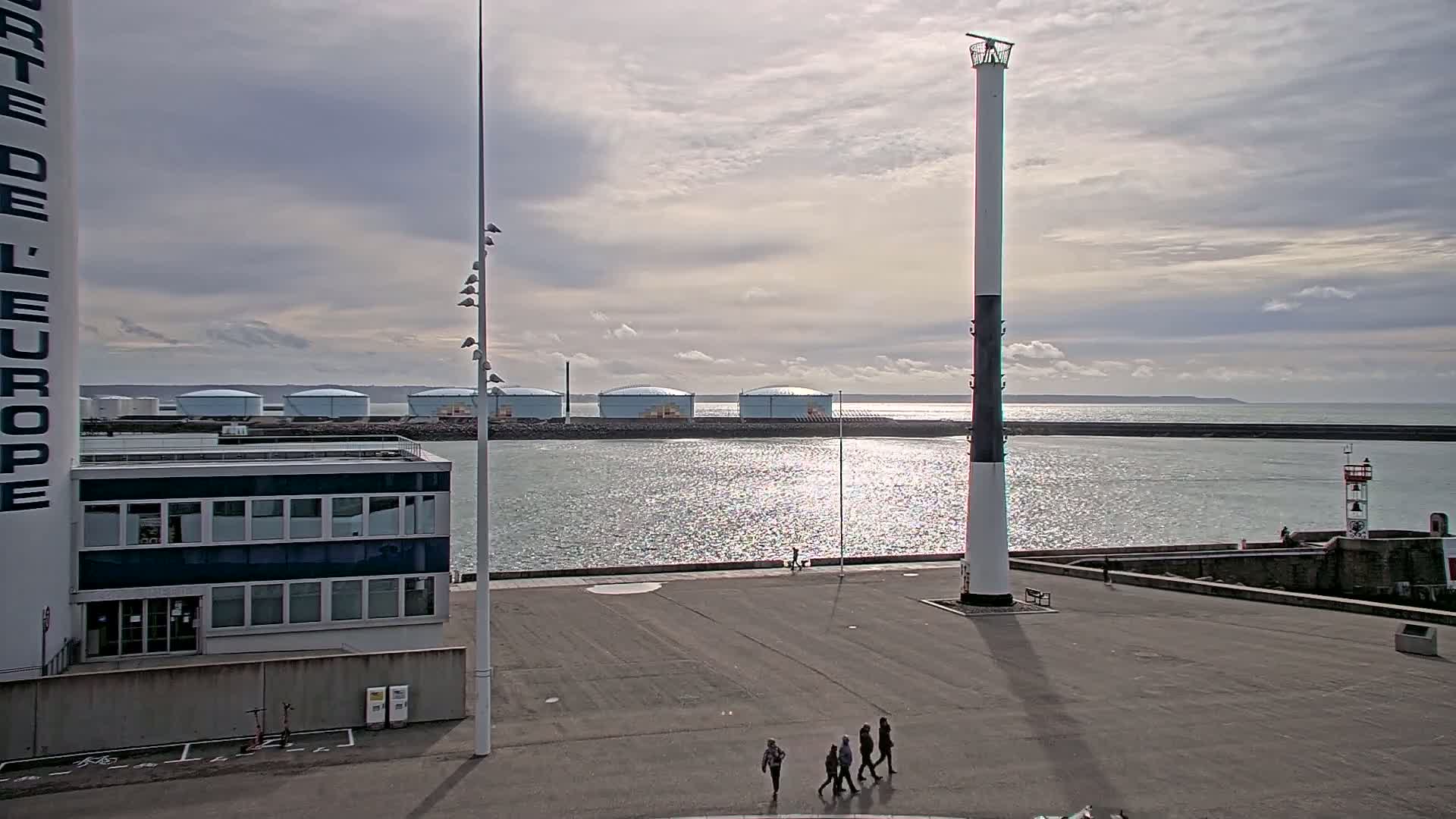 A mostly sunny day shows a harbor scene with a tall, slender lighthouse, several people walking, and a row of large storage tanks in the distance.