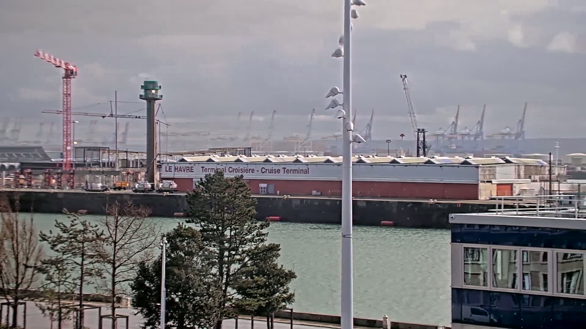A cruise terminal sits on a body of water under an overcast sky, with several cranes and other port infrastructure in the background.