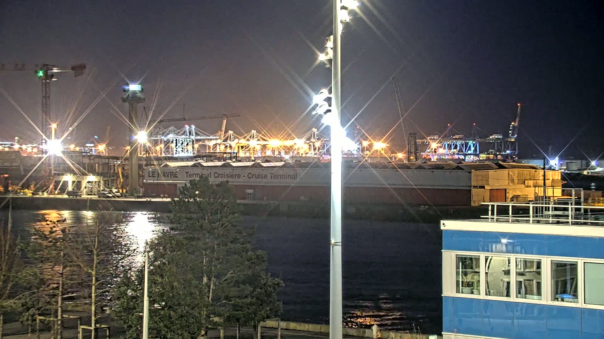 A brightly lit industrial port at night, viewed across a dark body of water, with several cranes and buildings visible.