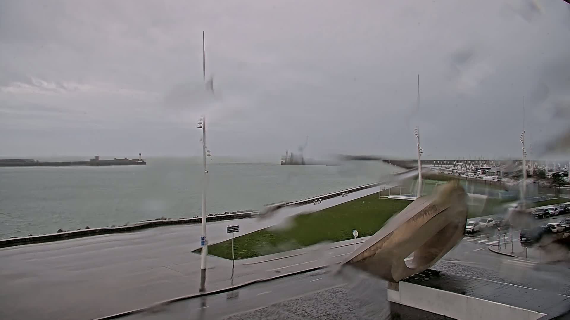 A rain-swept coastal scene shows a harbor with boats, a roadway, and a modern sculpture, all under a gray, overcast sky.