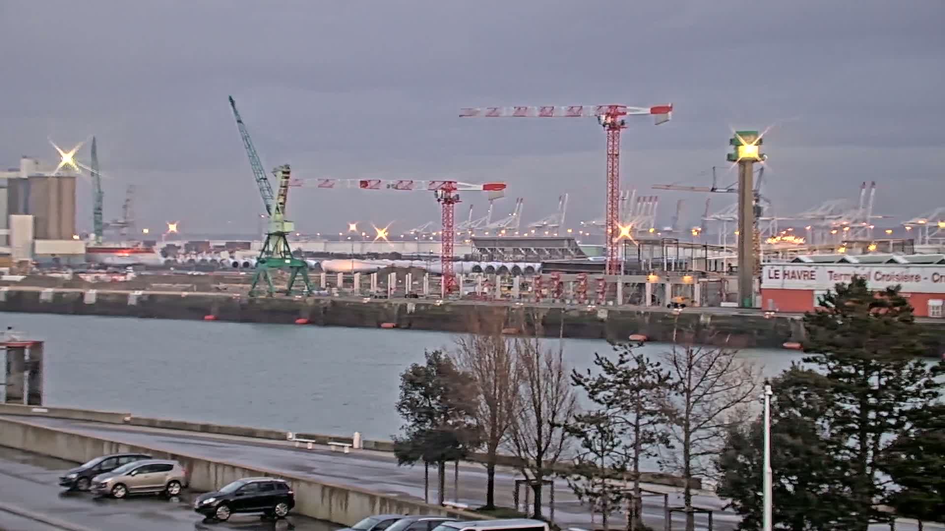 A harbor scene on an overcast day shows several cranes and industrial buildings along a waterfront, with cars parked along a nearby road and bare trees in the foreground.