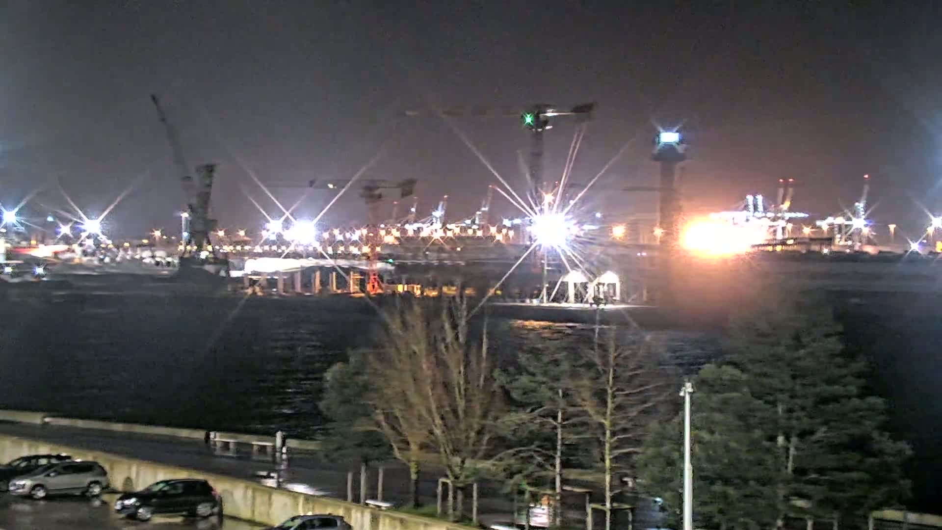 A brightly lit industrial port at night, seen from across a dark body of water, with bare trees and parked cars in the foreground under a dark sky.