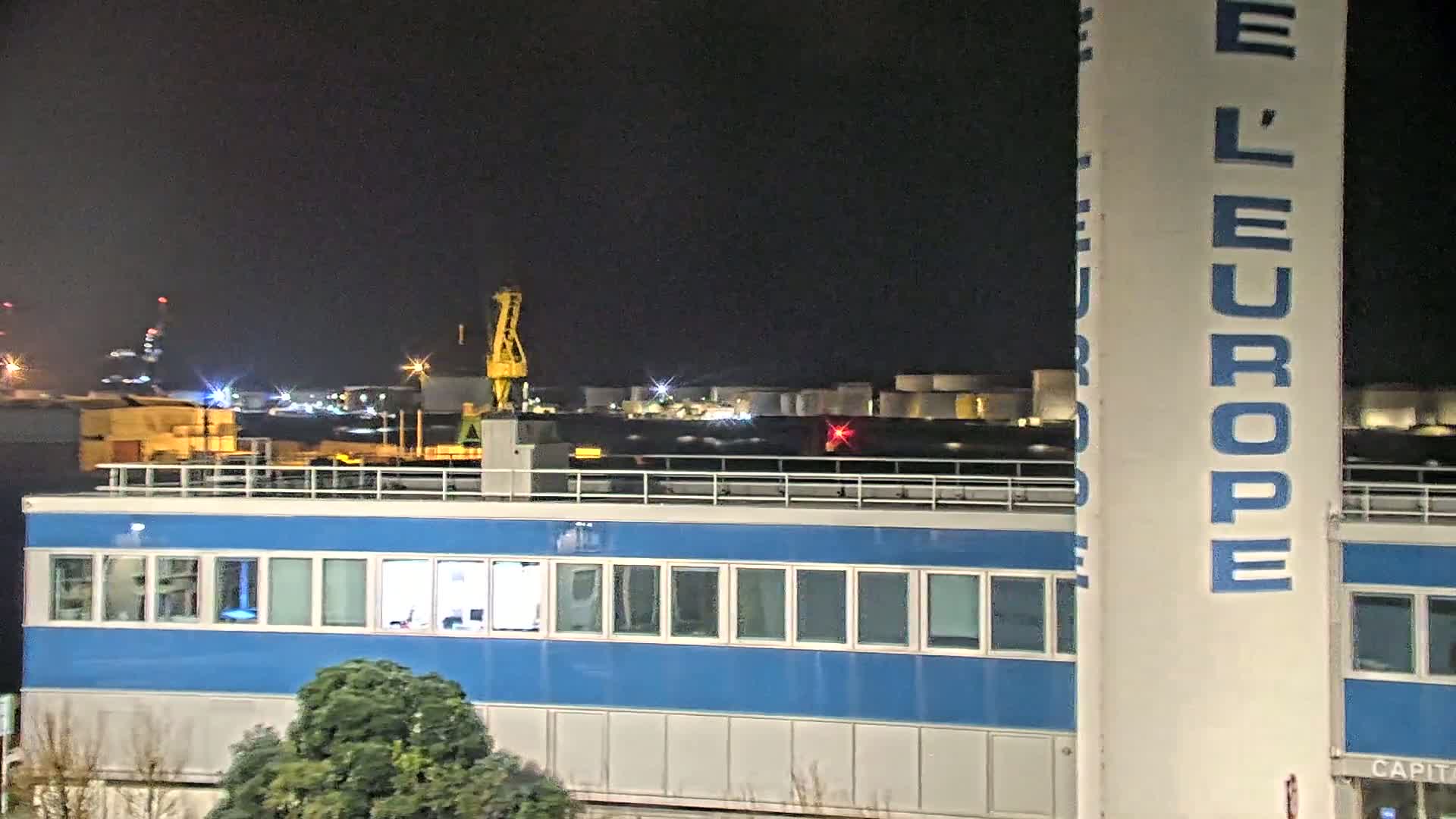 A blue building with many windows is seen at night, with a crane and industrial area in the background under a dark sky.