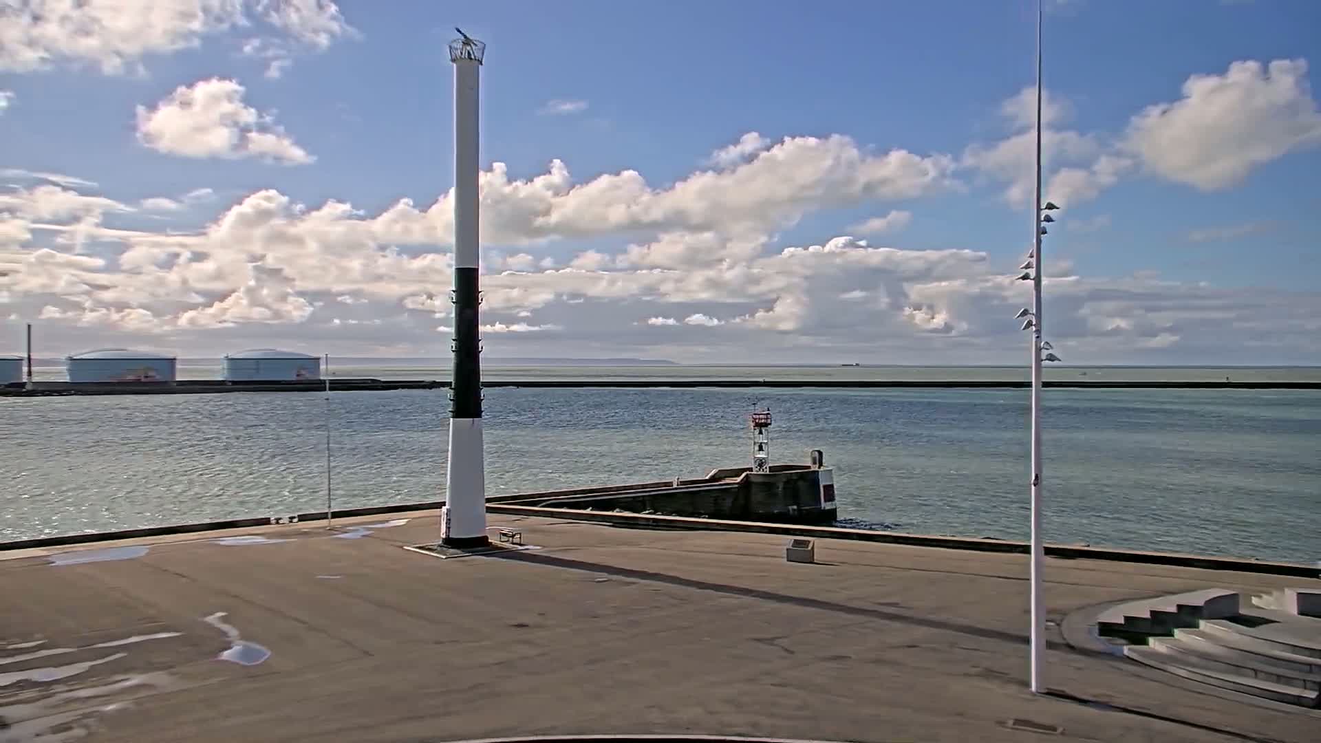 A tall, black and white lighthouse stands on a paved area overlooking a calm body of water under a partly cloudy sky, with a smaller lighthouse and several storage tanks visible in the distance.