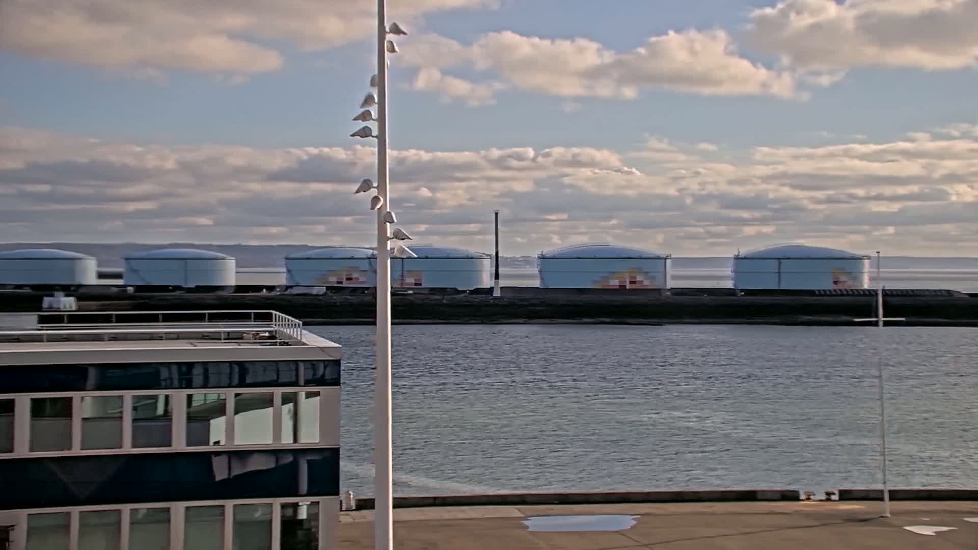 A partly cloudy sky overlooks a calm body of water with several large, light-blue storage tanks on a distant shore and a modern building in the foreground.