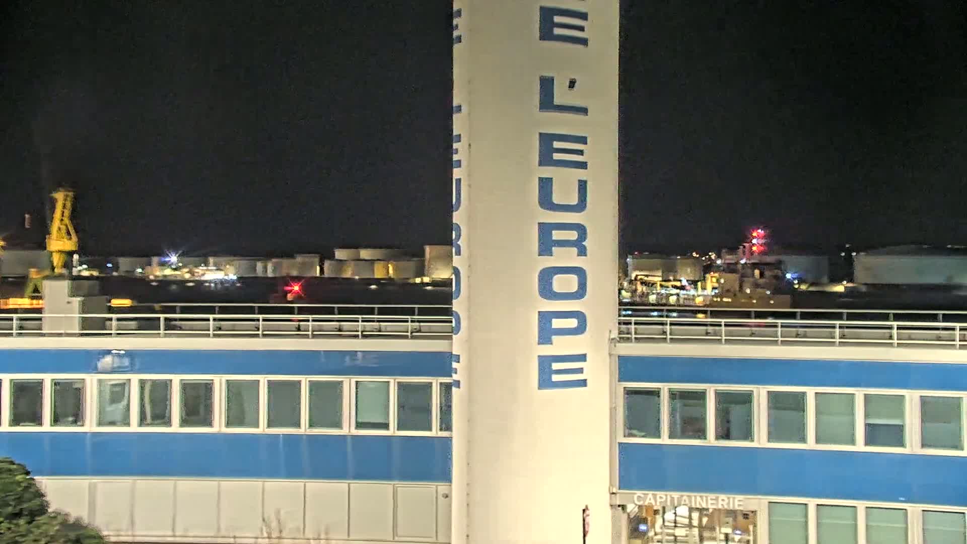 A nighttime view of a port, featuring a tall, white structure with blue lettering, a blue building with many windows, and industrial buildings and a crane in the background under a dark sky.