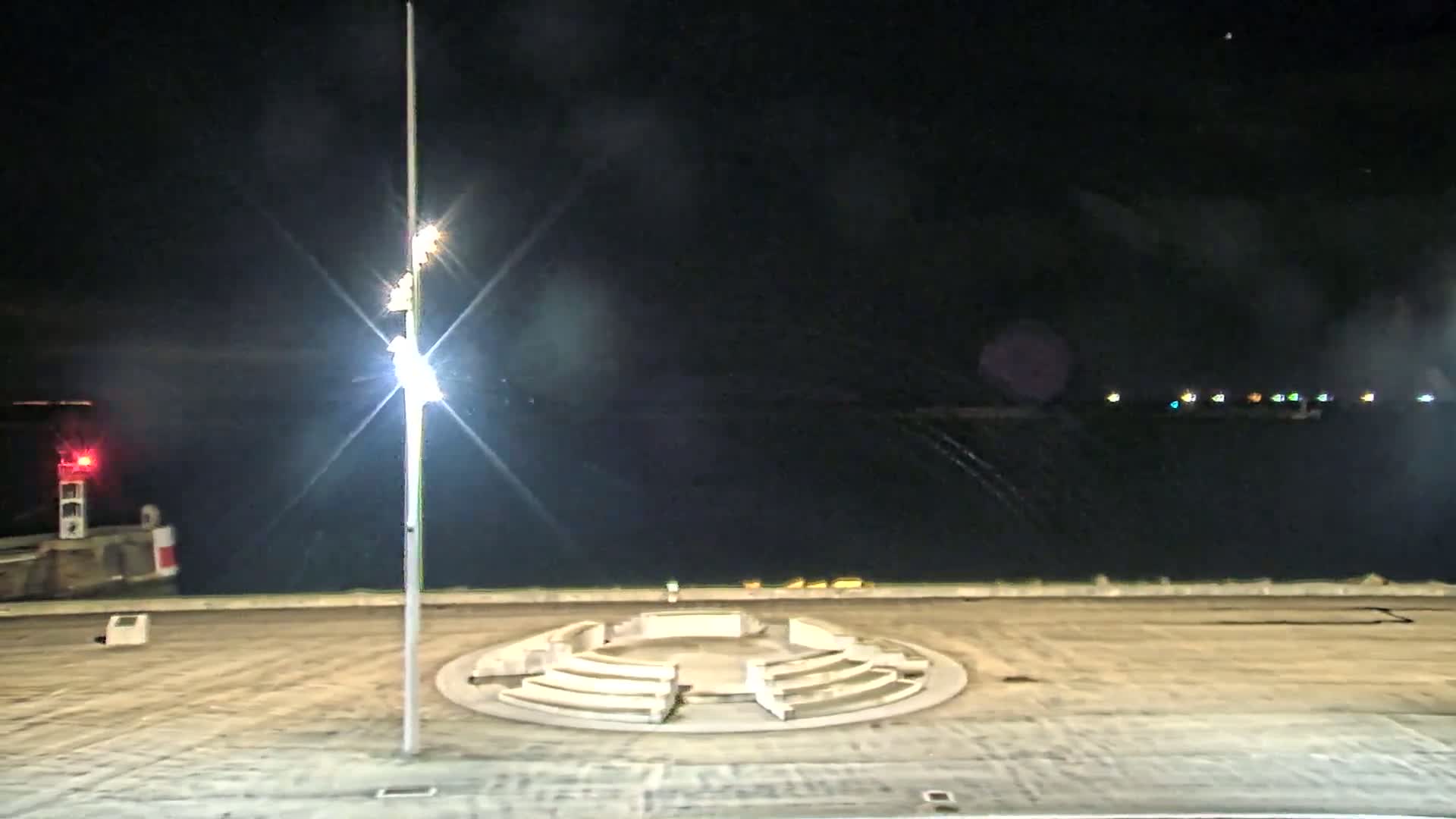 A nighttime view of a circular amphitheater-like structure on a waterfront, illuminated by a bright lamppost under a mostly clear sky.