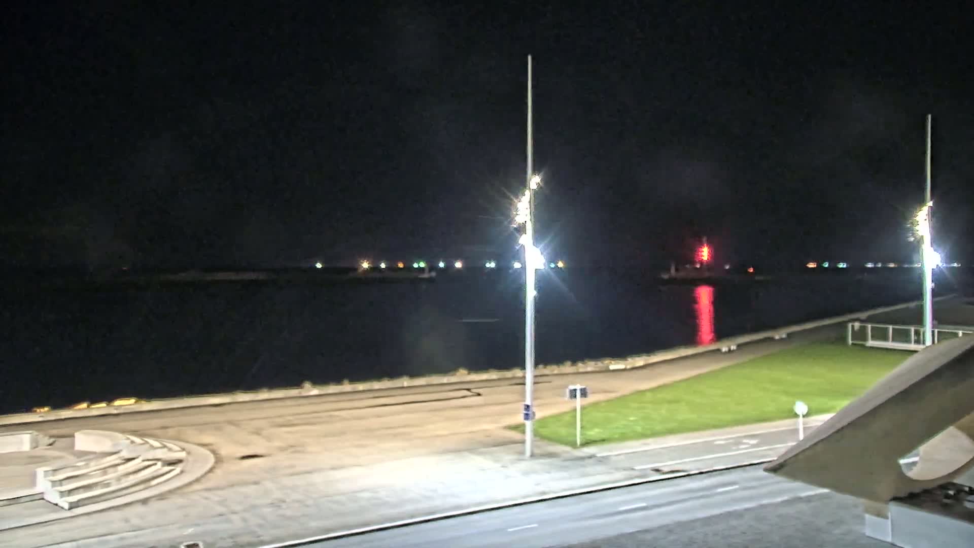 A nighttime view of a calm body of water with a paved waterfront area, grassy lawns, and tall light poles illuminating the scene.