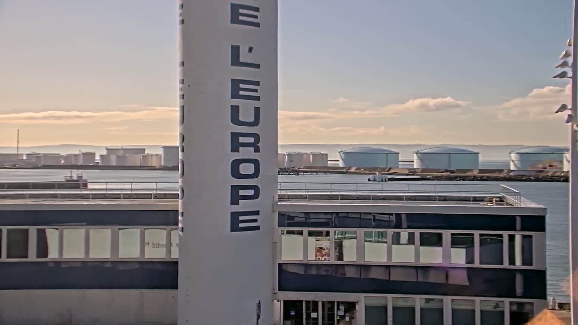 A tall, light-grey building with large windows is in the foreground, partially obscuring a view of a calm body of water with several large, light-grey storage tanks in the background under a partly cloudy sky.