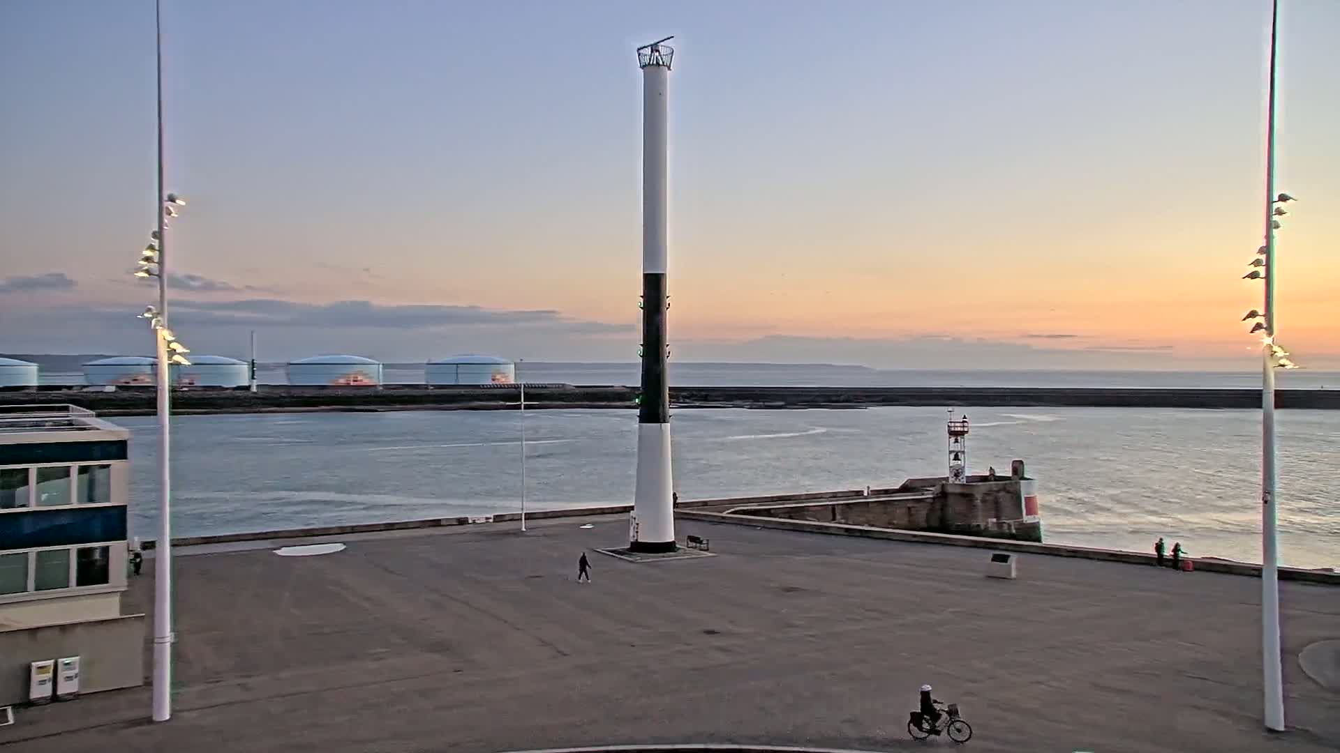 A tall, slender lighthouse stands in a harbor at sunset, with a calm body of water, several storage tanks in the background, and a person on a bicycle in the foreground.