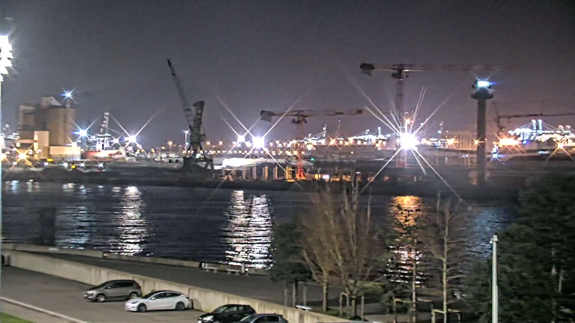 A nighttime view of a brightly lit harbor with cranes, industrial buildings, and calm water reflecting the lights, seen from across a waterfront area with parked cars and trees.
