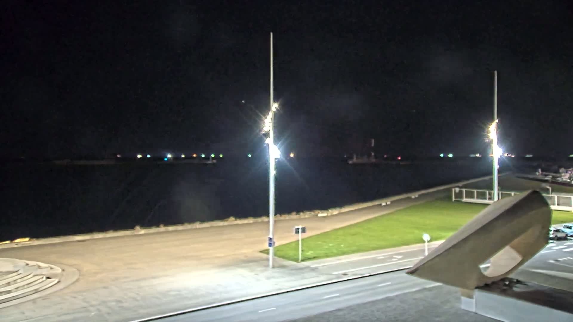 A nighttime view of a waterfront area with a calm body of water, illuminated by tall lights, and a grassy area next to a paved road.