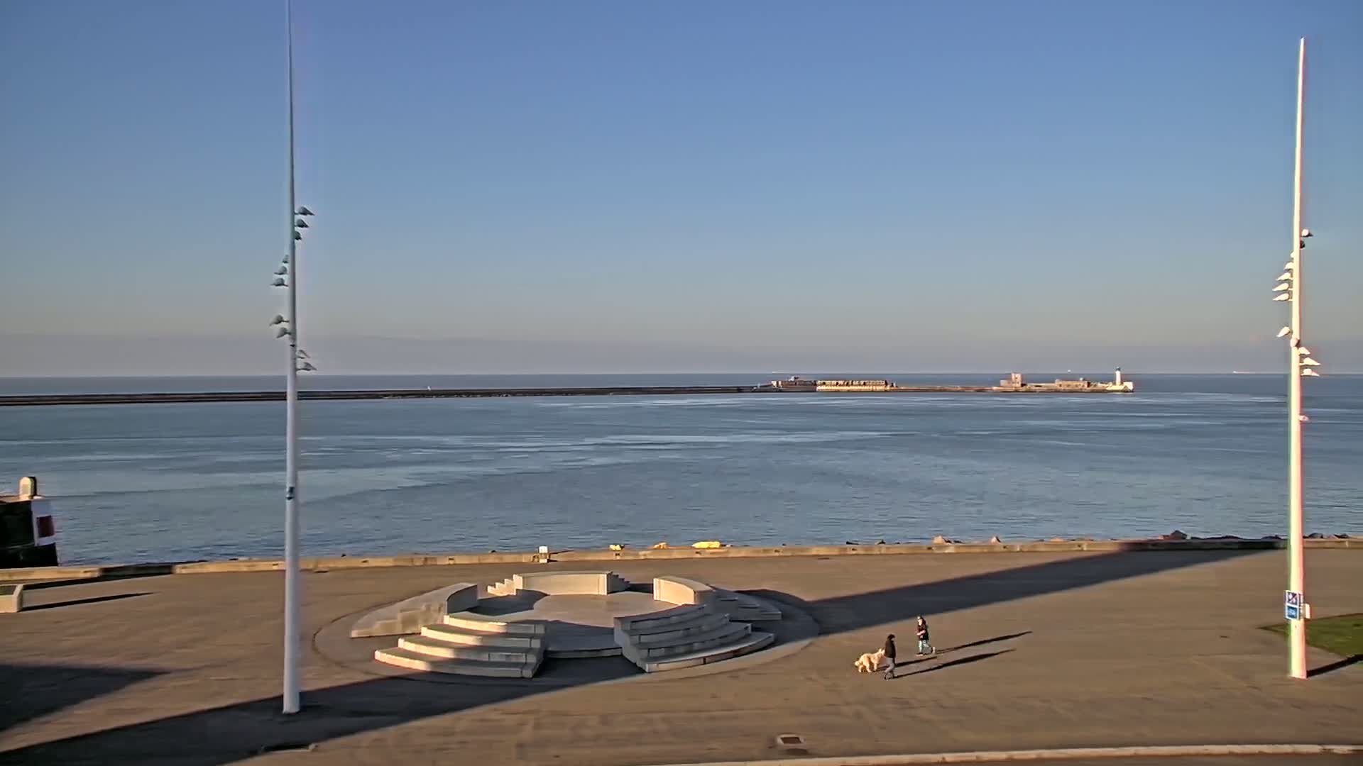 A paved seaside plaza with a circular amphitheater-like structure features two people walking a dog, overlooking calm water under a clear blue sky.
