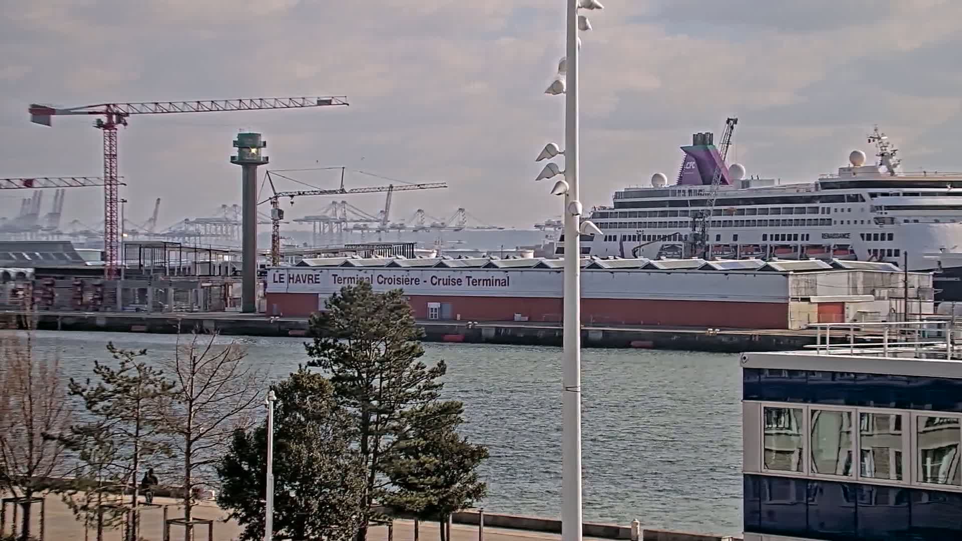 A cruise ship is docked at a port under a partly cloudy sky, with construction cranes and other port infrastructure visible in the background.