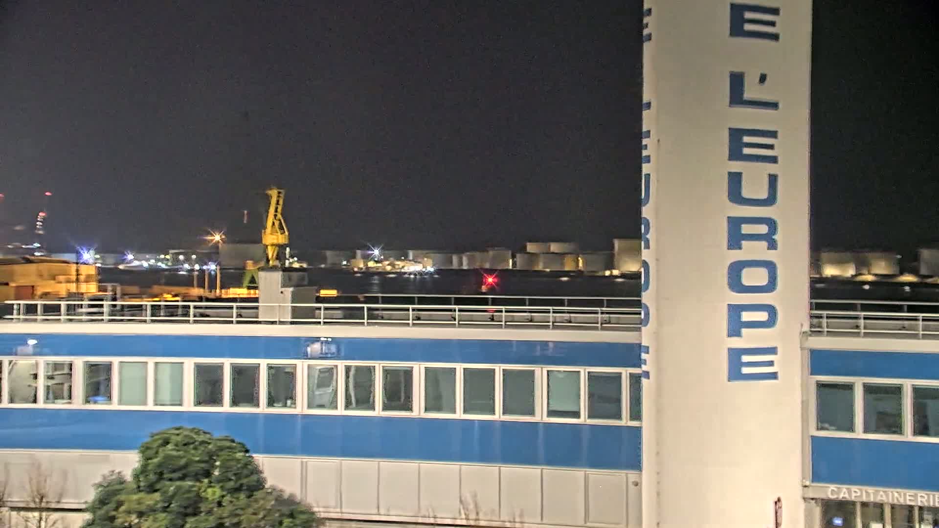 A nighttime view of a blue building with numerous windows, a tall white tower with blue lettering, a crane, and a body of water with industrial structures and lights in the background.