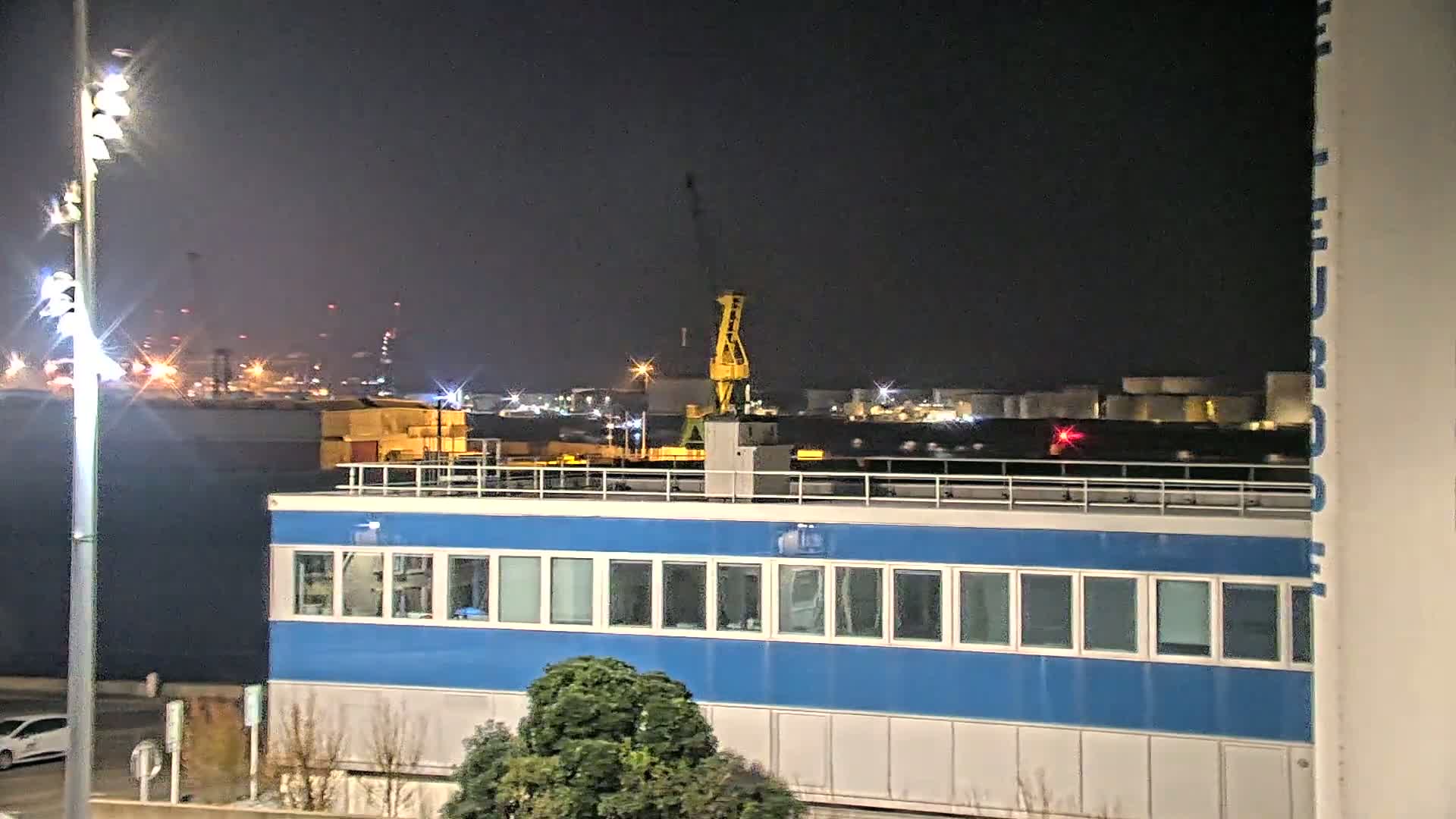 At night, a blue building with many windows sits in the foreground, with a distant industrial area illuminated under a dark sky.