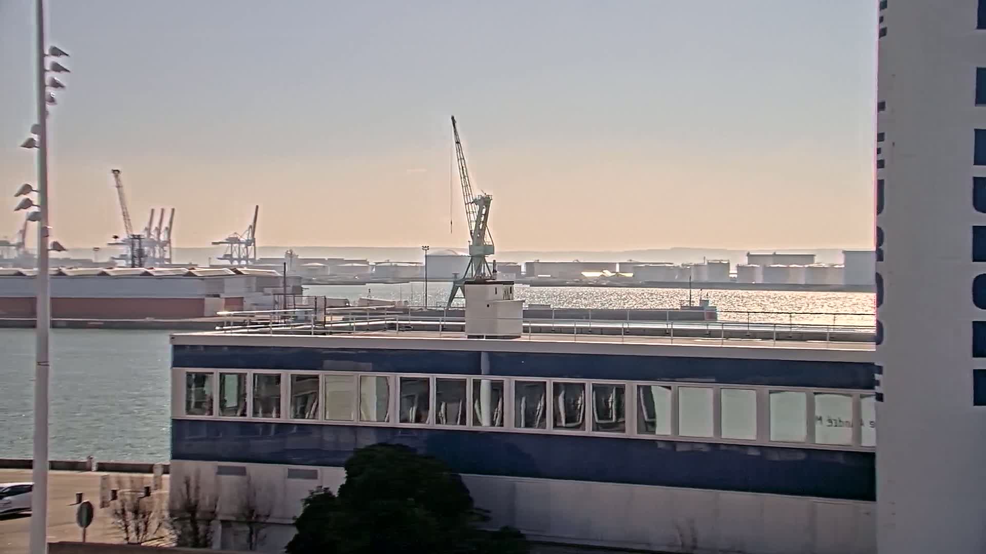 A blue building with many windows sits on a waterfront, overlooking a harbor with several cranes and large industrial storage tanks under a bright, sunny sky.