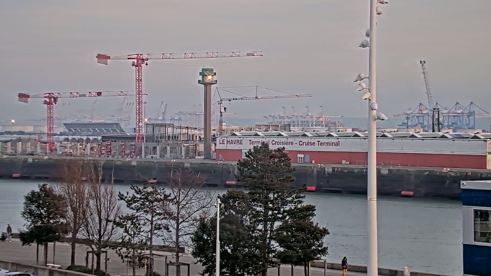 A partially cloudy day reveals a harbor scene with construction cranes near a cruise terminal building and numerous cargo ships in the distance.