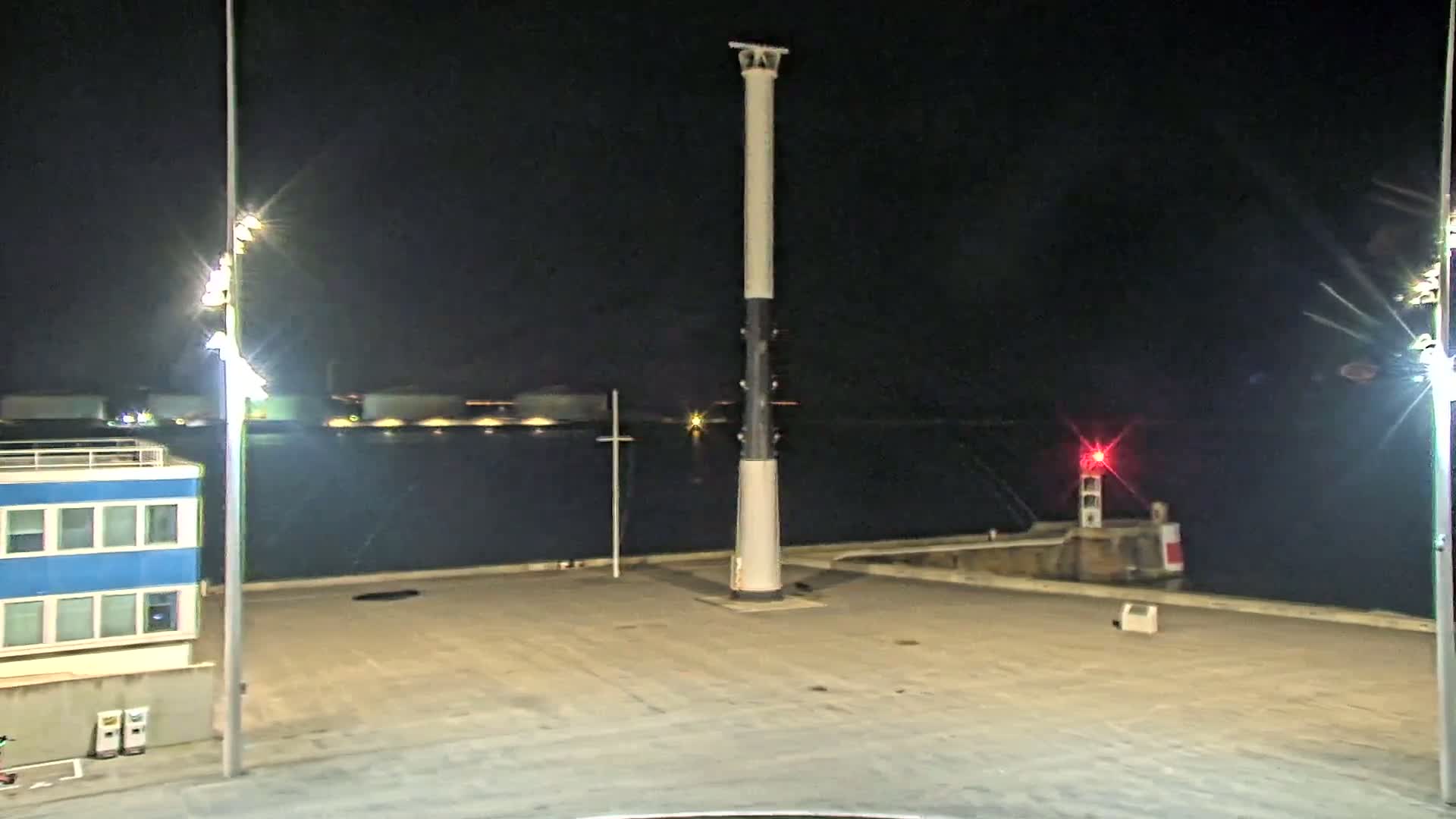 A tall, slender light post stands in a dark, empty harbor area at night, with a smaller red light visible in the distance and bright lights illuminating the scene.