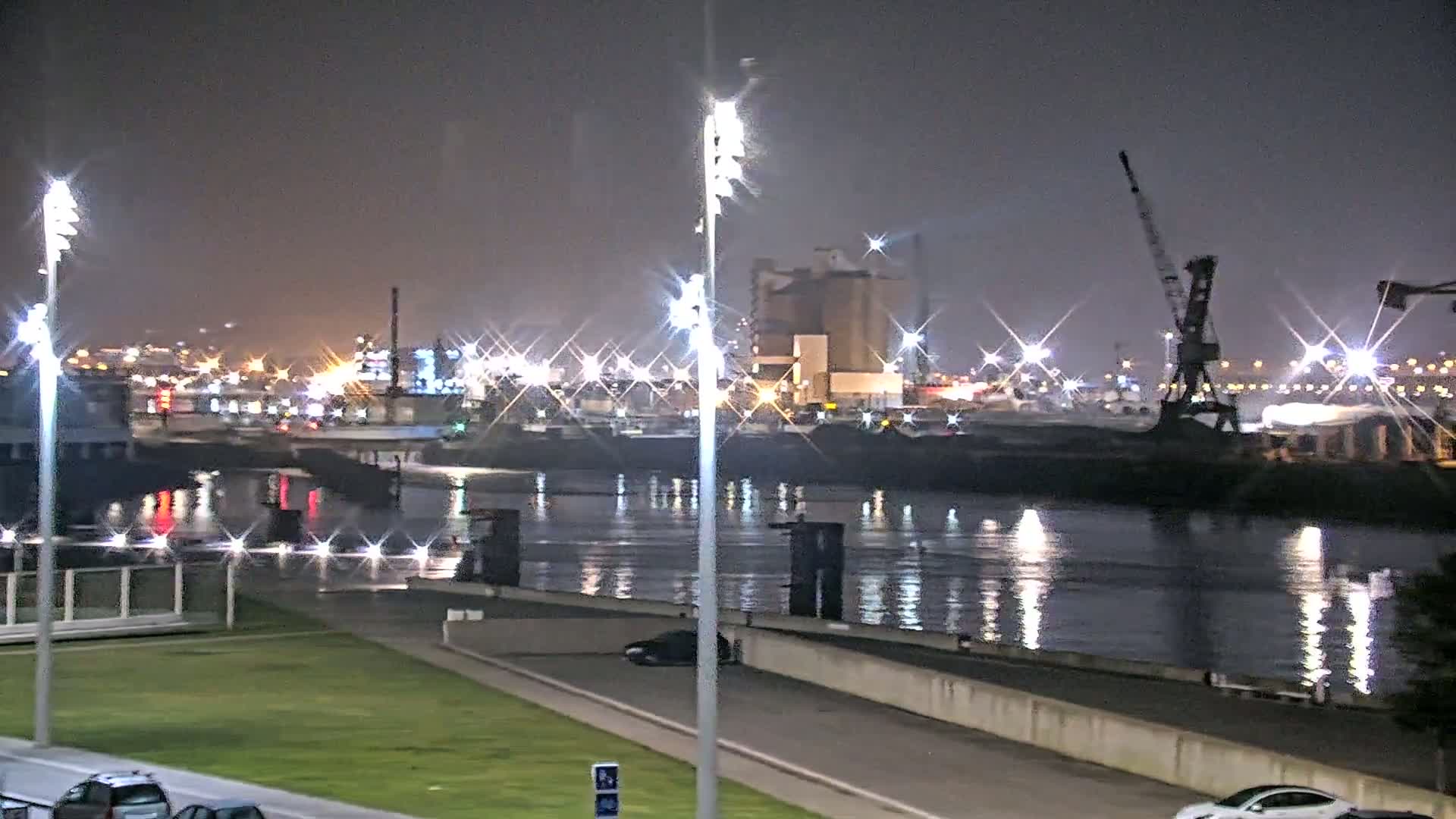 At night, a calm body of water reflects the lights of an industrial waterfront area with cranes and buildings.