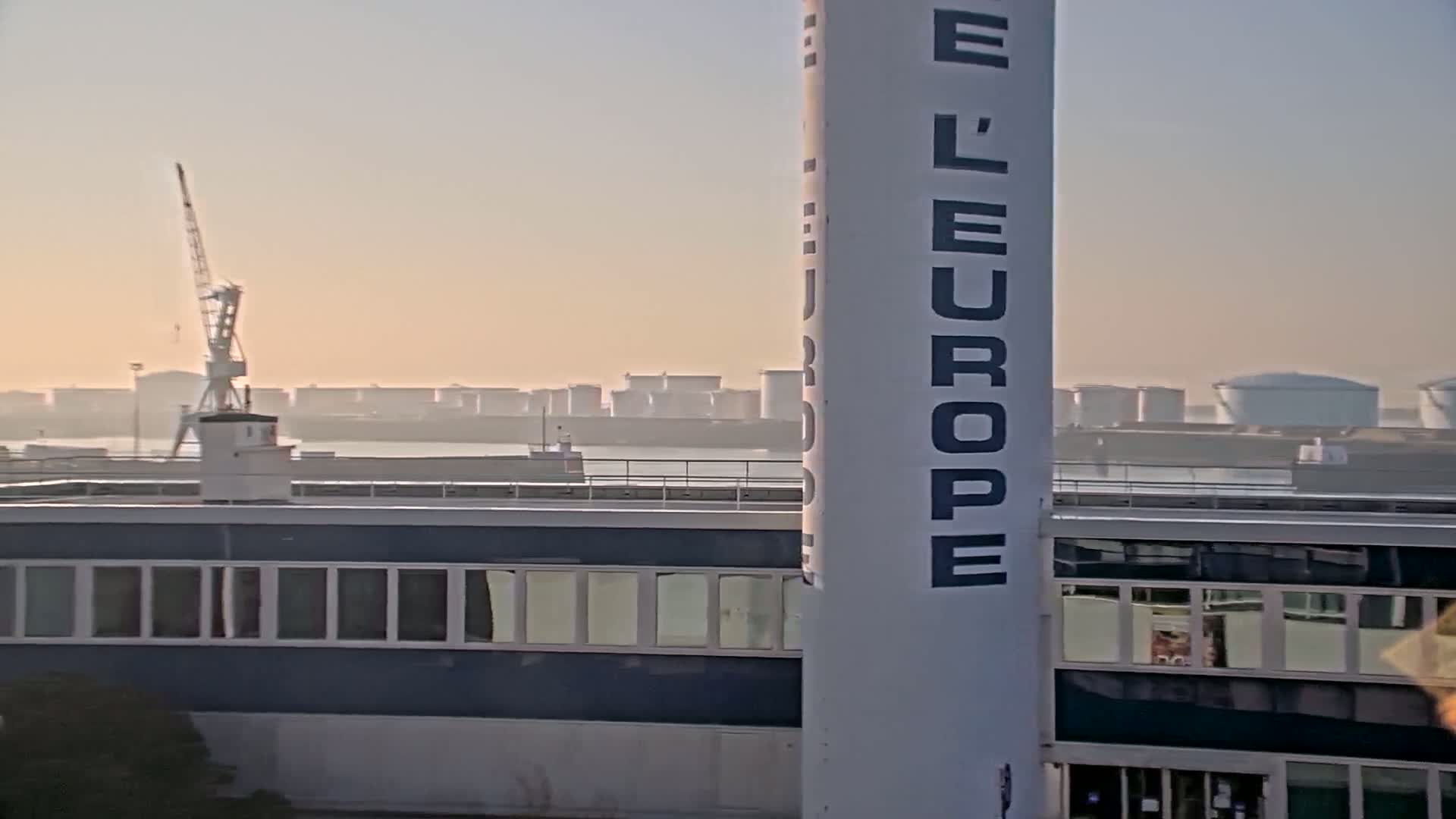 A hazy sunrise illuminates a harbor scene featuring a tall structure with "L'EUROPE" written vertically, a building with numerous windows, and distant industrial structures and a crane.
