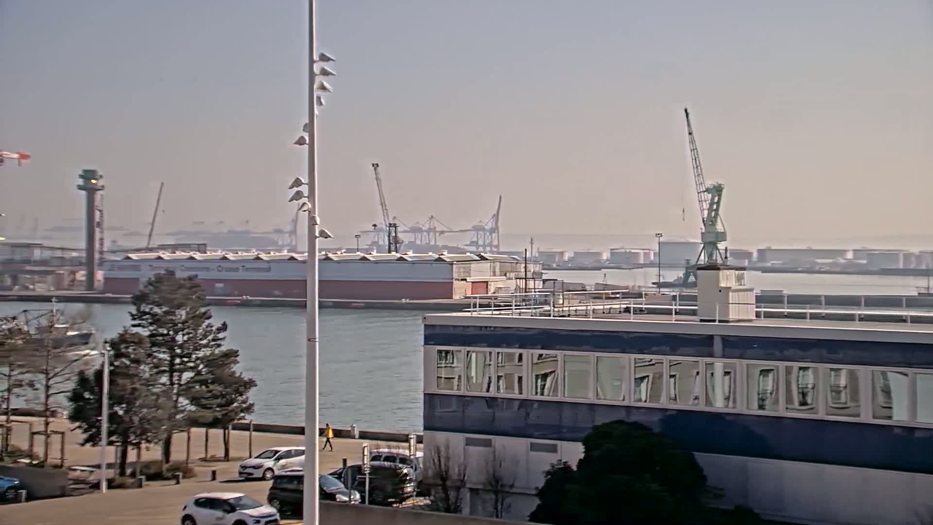 A hazy day shows a harbor with several cranes, industrial buildings, and parked cars near a body of water.