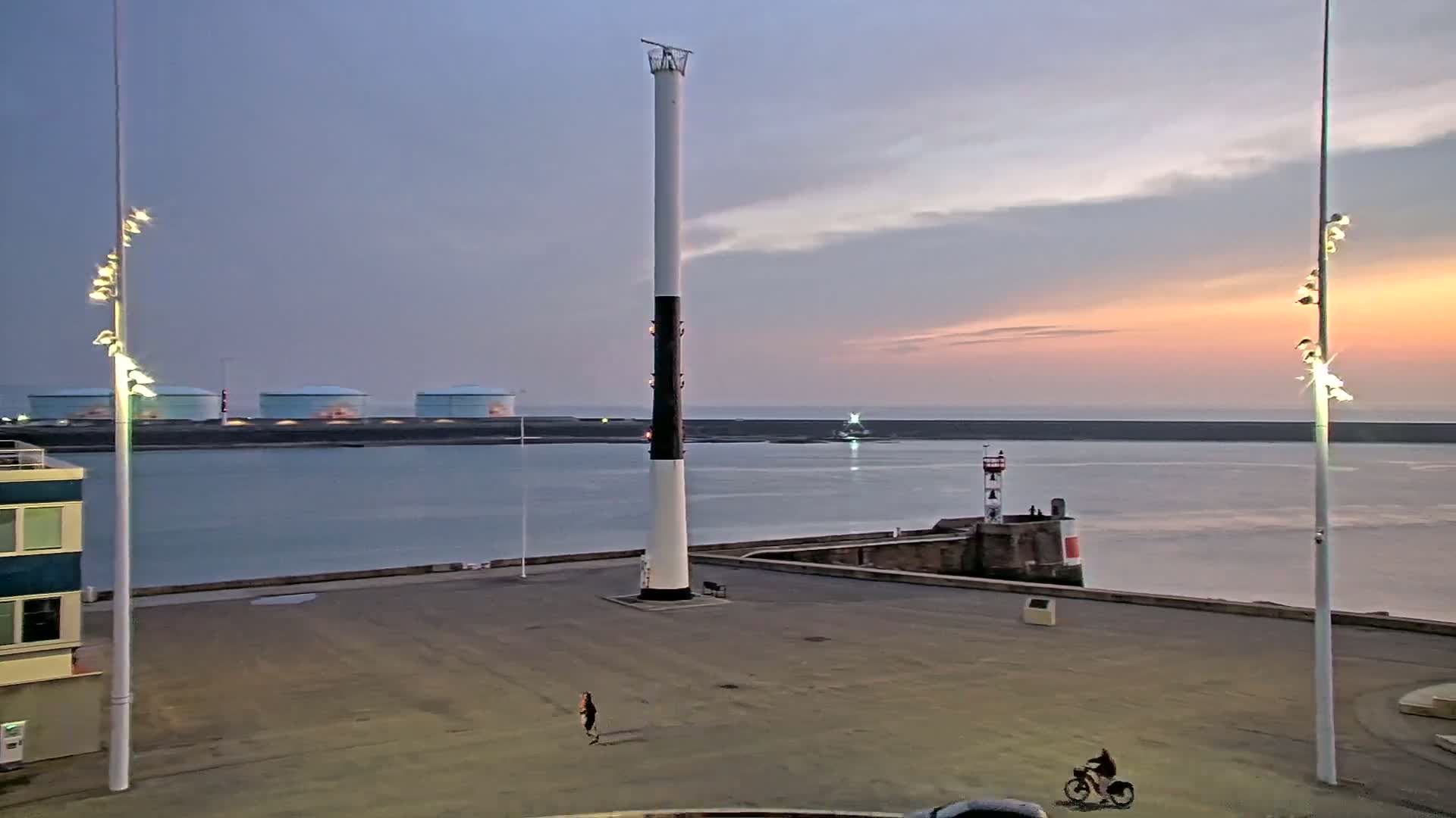A tall, black-and-white striped tower stands in a harbor at sunset, with a person jogging and another riding a bicycle in the foreground.