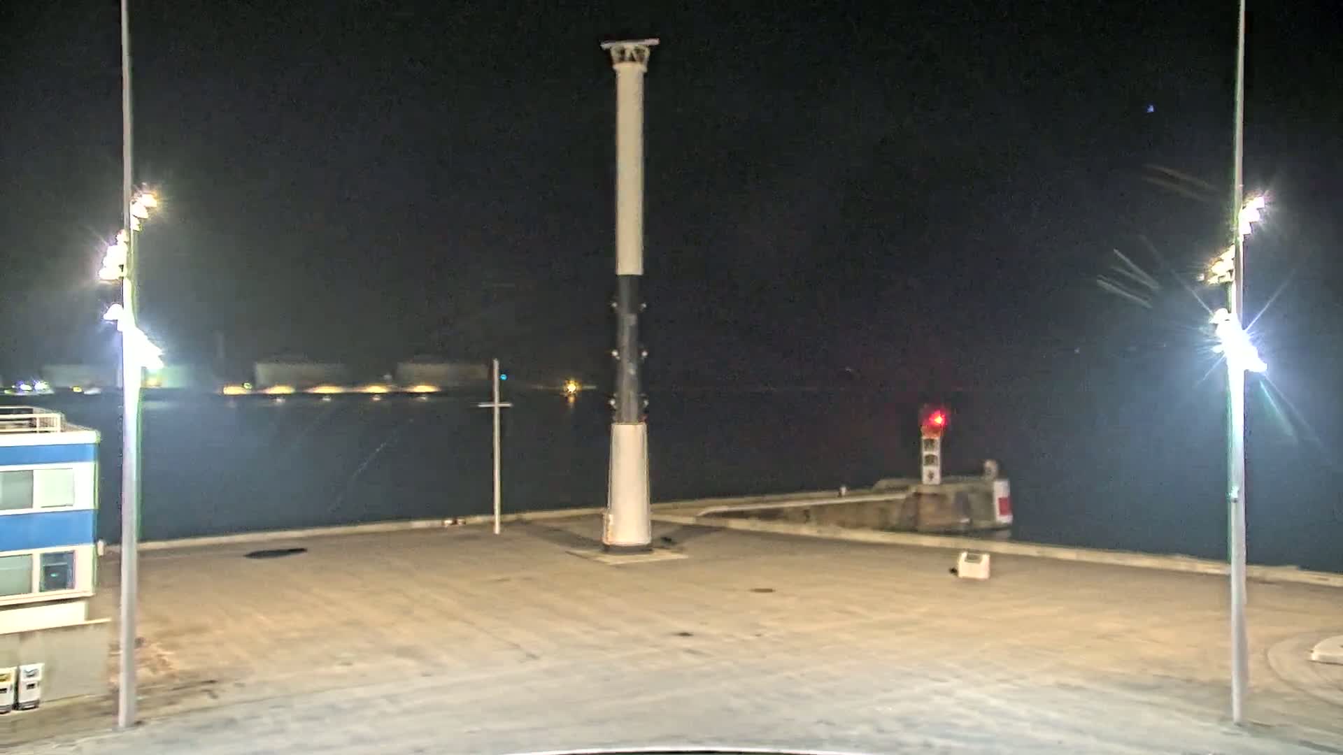At night, a tall, mostly white pillar stands in a paved area next to a body of dark water, with bright lights illuminating the scene and a smaller red light visible in the distance.